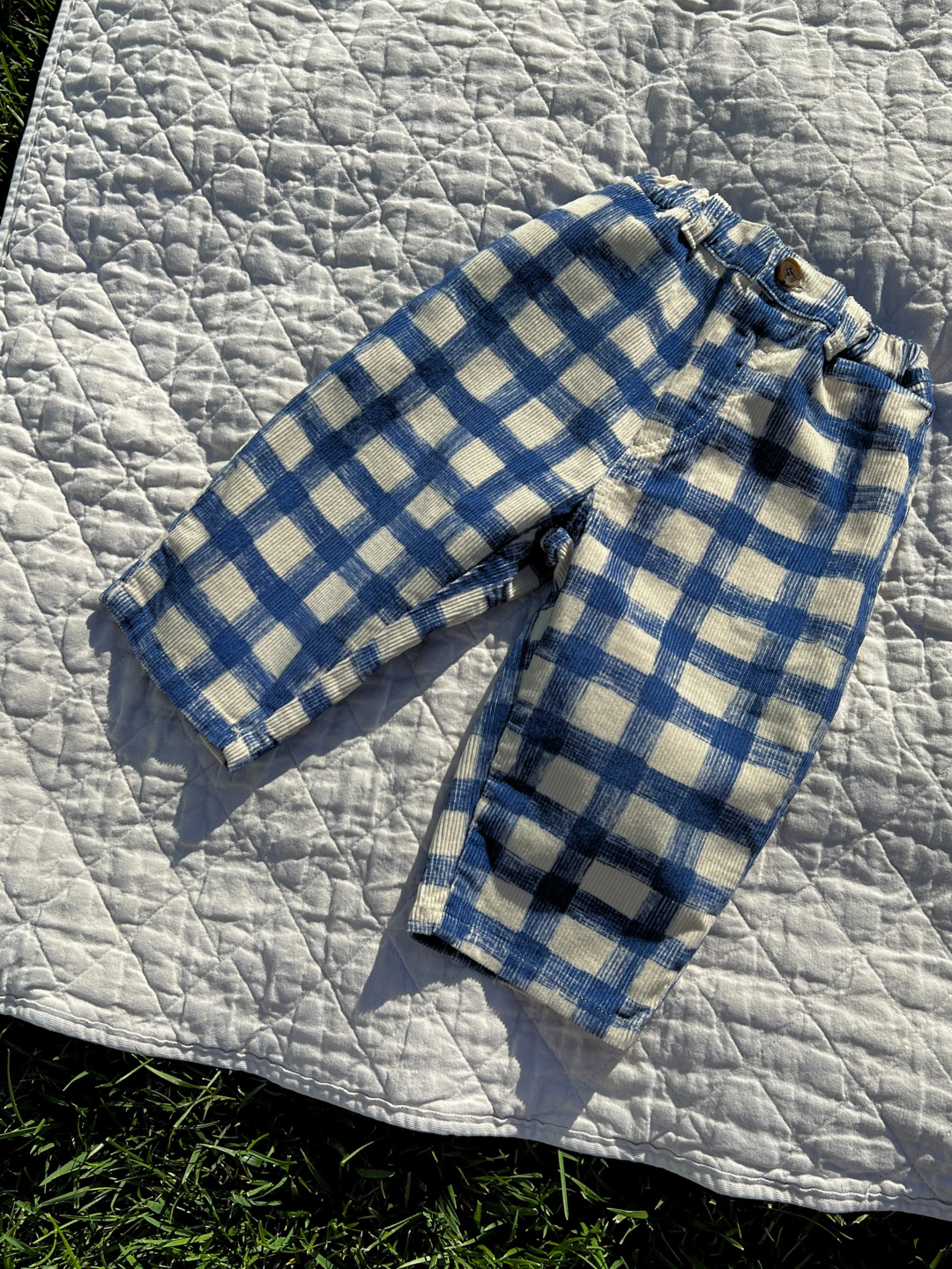 Front view of kids corduroy trousers in white with a large blue grid check pattern. Pants are shown on a white canvas blanket on grass.
