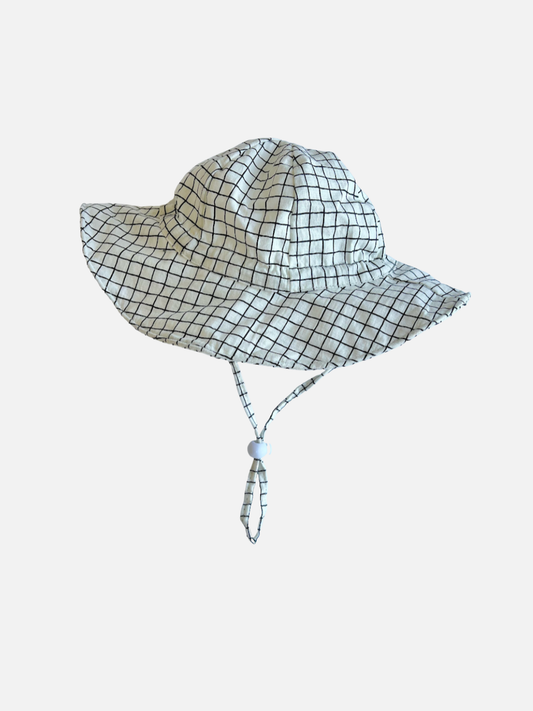 Second image of White | A baby wearing the Off the Grid Hat in white with a black grid check pattern. The hat has a flexible brim and a chinstrap, and the baby wears a blue top and sits in the arms of an adult in a grey tshirt, with a street scene background.