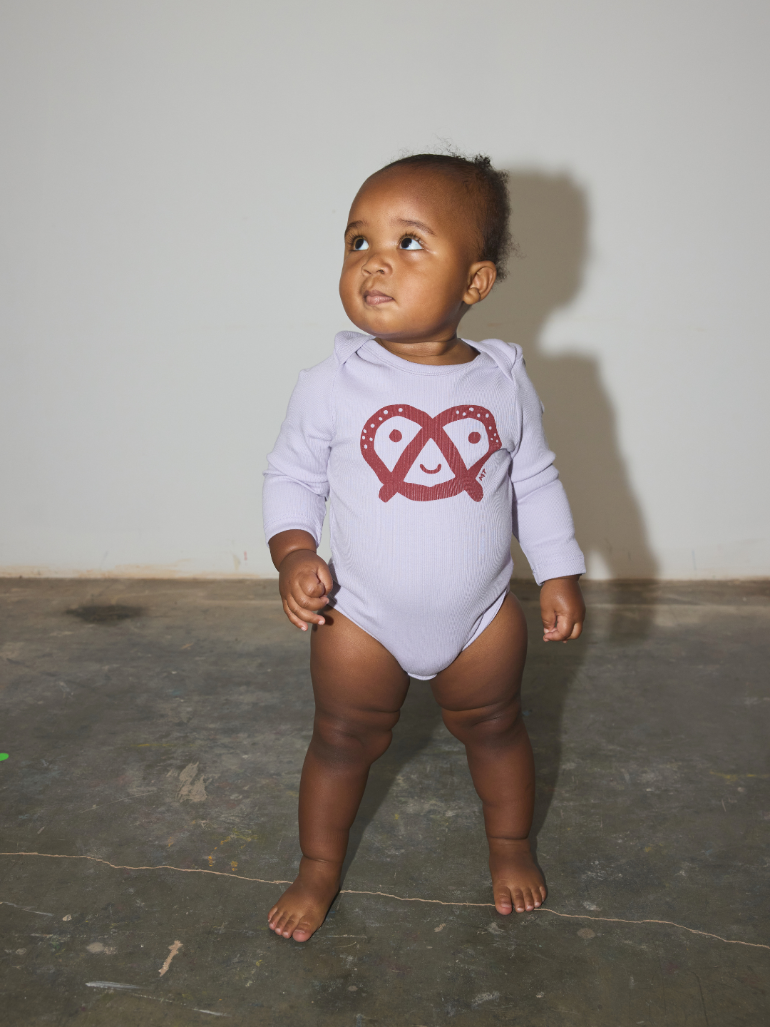 A baby standing barefoot on a concrete floor against a plain wall, wearing a light purplelong-sleeve bodysuit with a red pretzel graphic on the front.