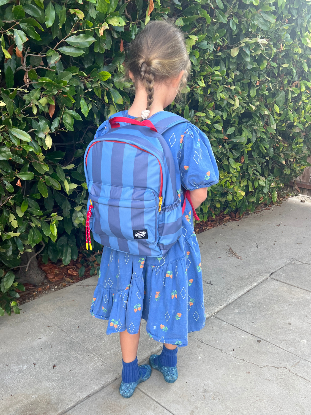 A young girl with braided hair is shown from behind, wearing a blue patterned dress and a blue striped backpack with red accents, standing on a concrete sidewalk next to a green hedge.