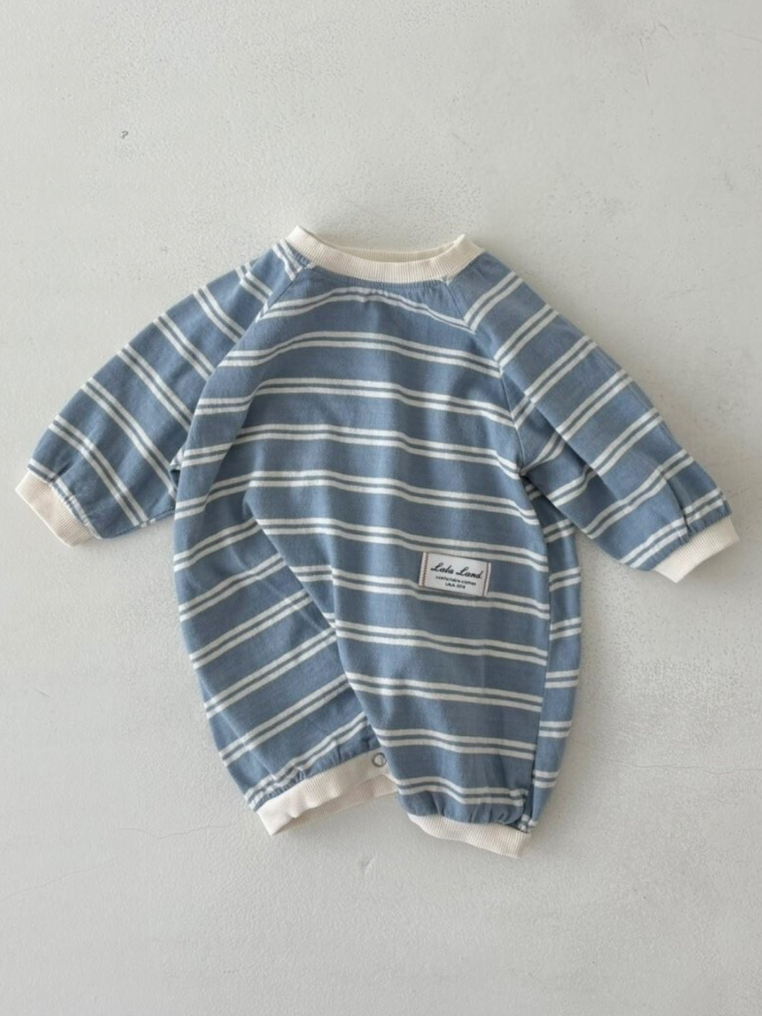 The SLOUCHY STRIPE ROMPER is a soft cotton jersey blue and white striped romper with long sleeves, cream cuffs, and a patch label—perfect for your little one from a favorite Korean baby brand.