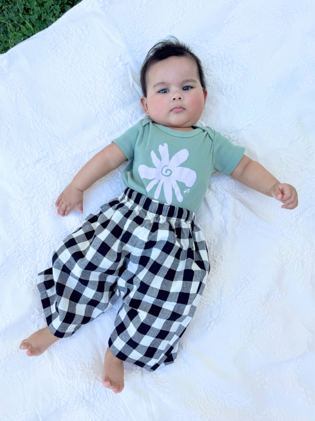 Green | A baby lies outdoors on a white blanket, wearing the FLOWER SHORT SLEEVE ONESIE in green ribbed cotton with a white flower graphic, offering comfort and style that's perfect for easy diaper changes.