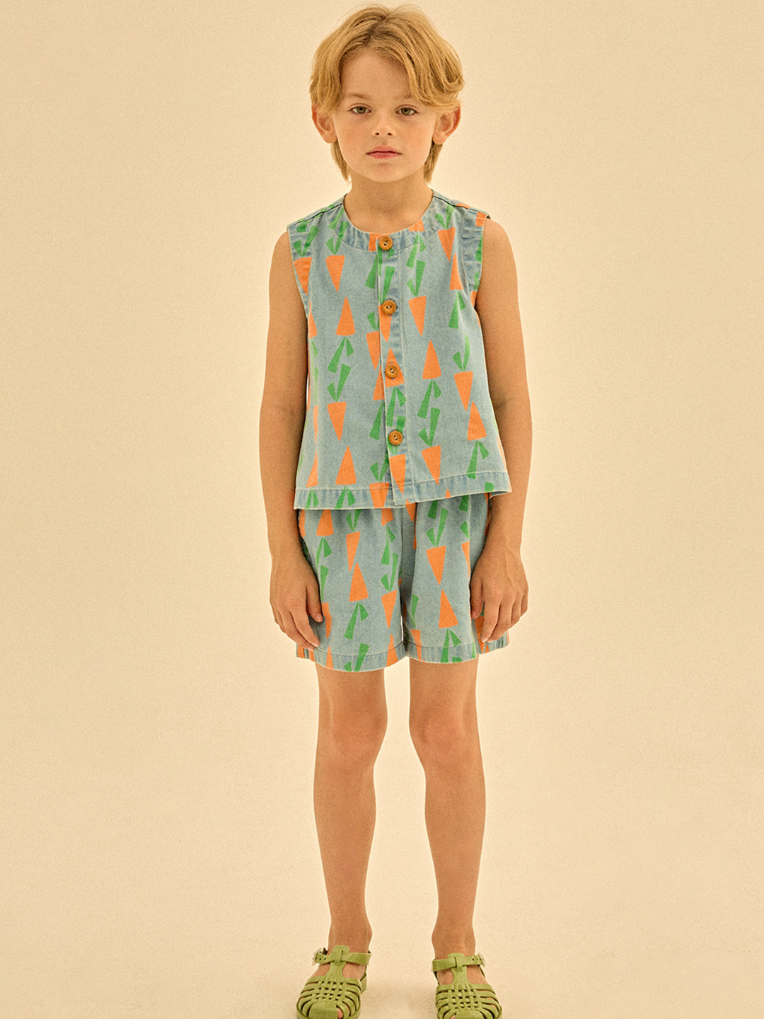 A child in a sleeveless, button-down outfit featuring a tree pattern stands against a plain background, wearing green sandals along with CARROT DENIM SHORTS.