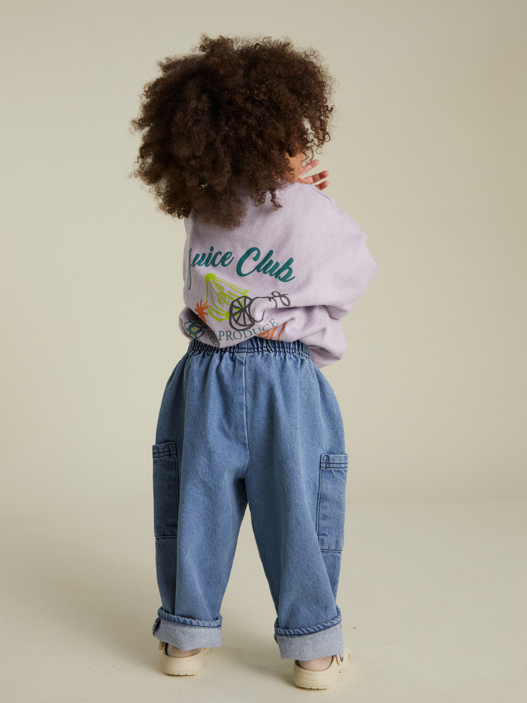 A young child with curly hair stands facing away, wearing a light purple sweatshirt with "Juice Club" on the back, EMERSON DENIM BABY TROUSER made from organic cotton, and beige shoes.