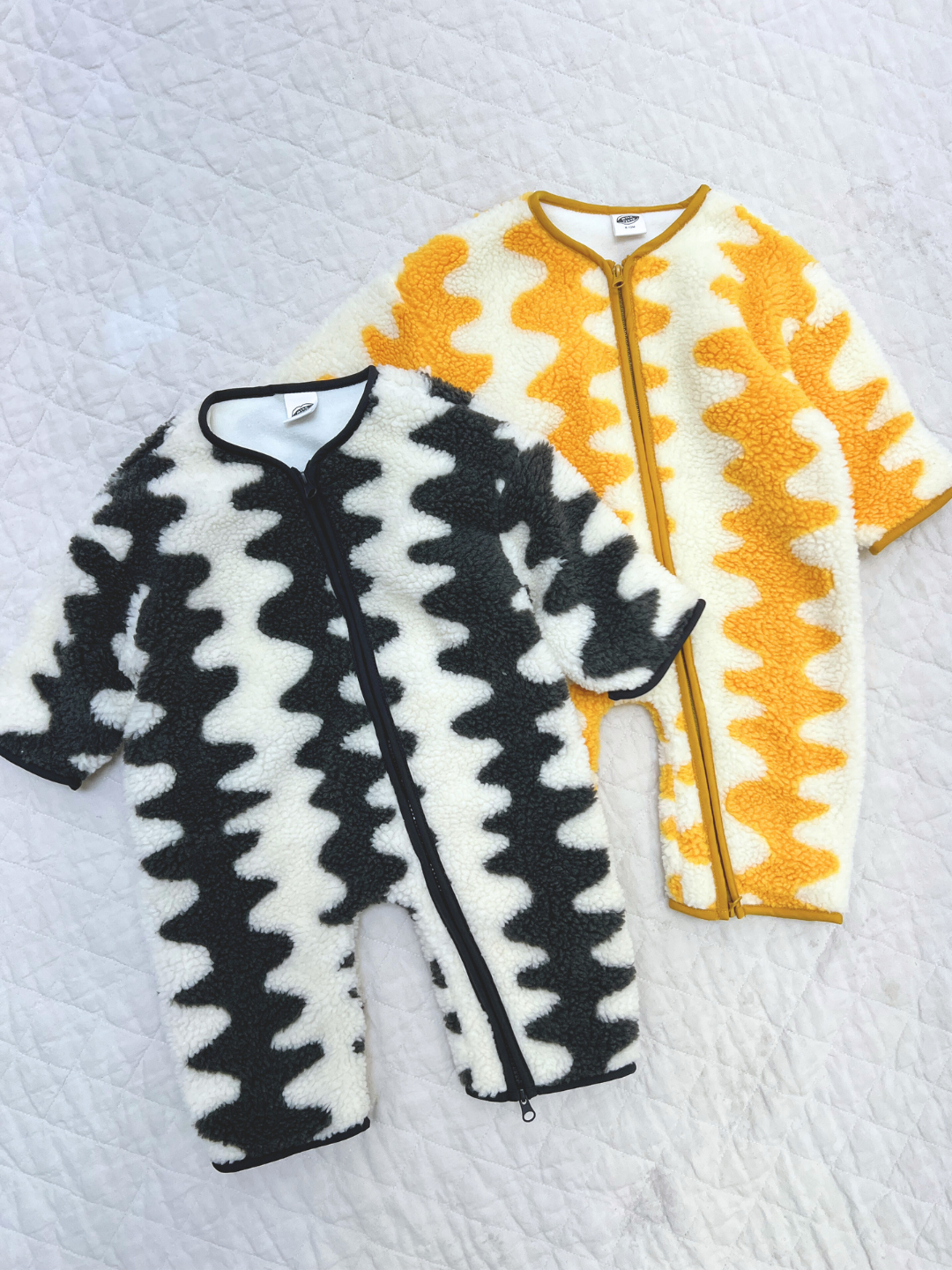 Yellow | Two WAVELENGTH SHERPA FLEECE STROLLER SUITS—one black and white, one yellow and white—feature cozy wavy zigzag patterns, making them perfect for layering and keeping baby warm in winter. Shown on a quilted white surface.