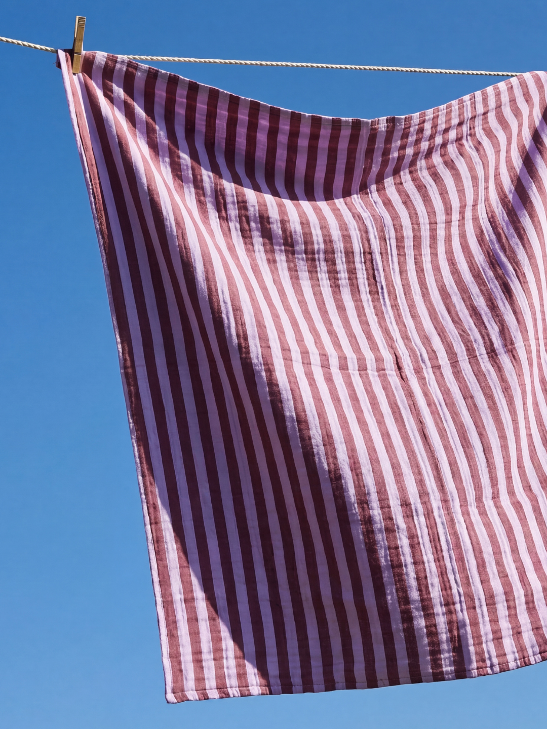 Purple Stripe | The organic cotton swaddle blanket in purple and maroon stripes is shown hanging on a washing line against blue sky.