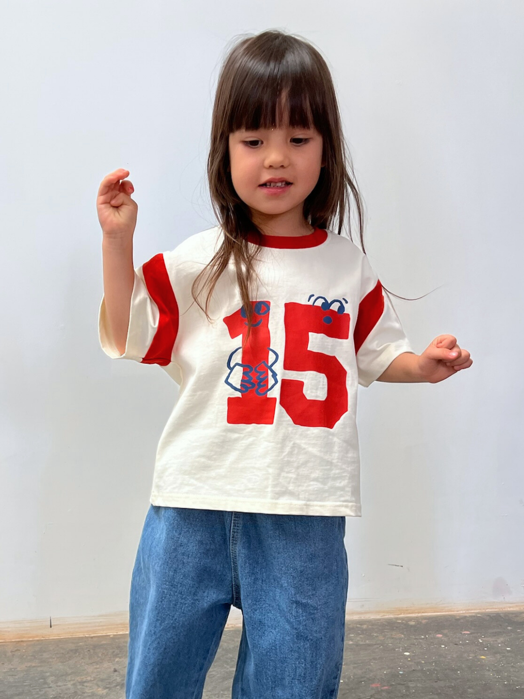Child wearing a white t-shirt with red numbers and blue pants against a plain background