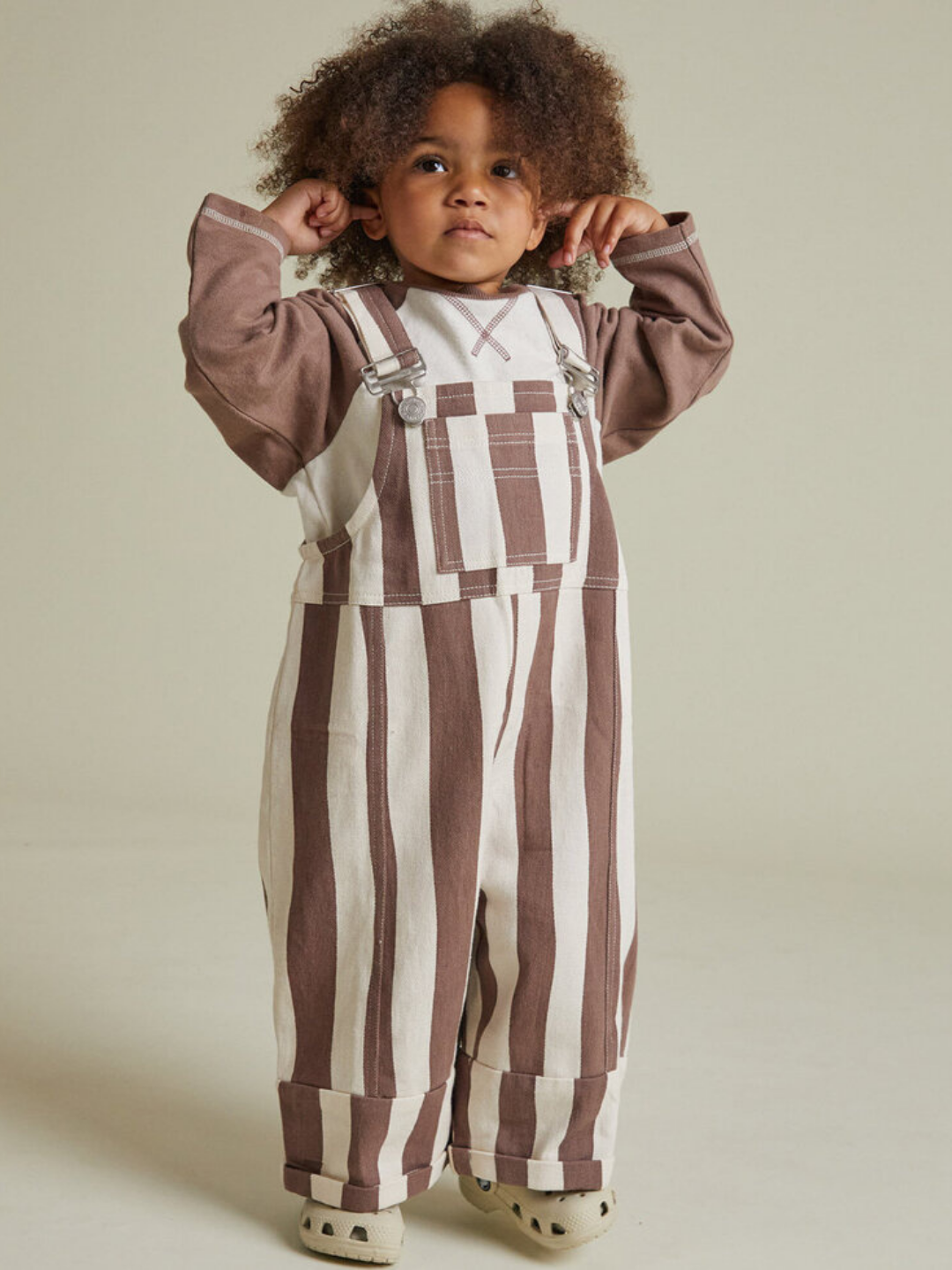 A young child indoors wears the BLYTHE CHOC STRIPE DUNGAREE, paired with a brown long-sleeve shirt and cream clogs, touching their ears.