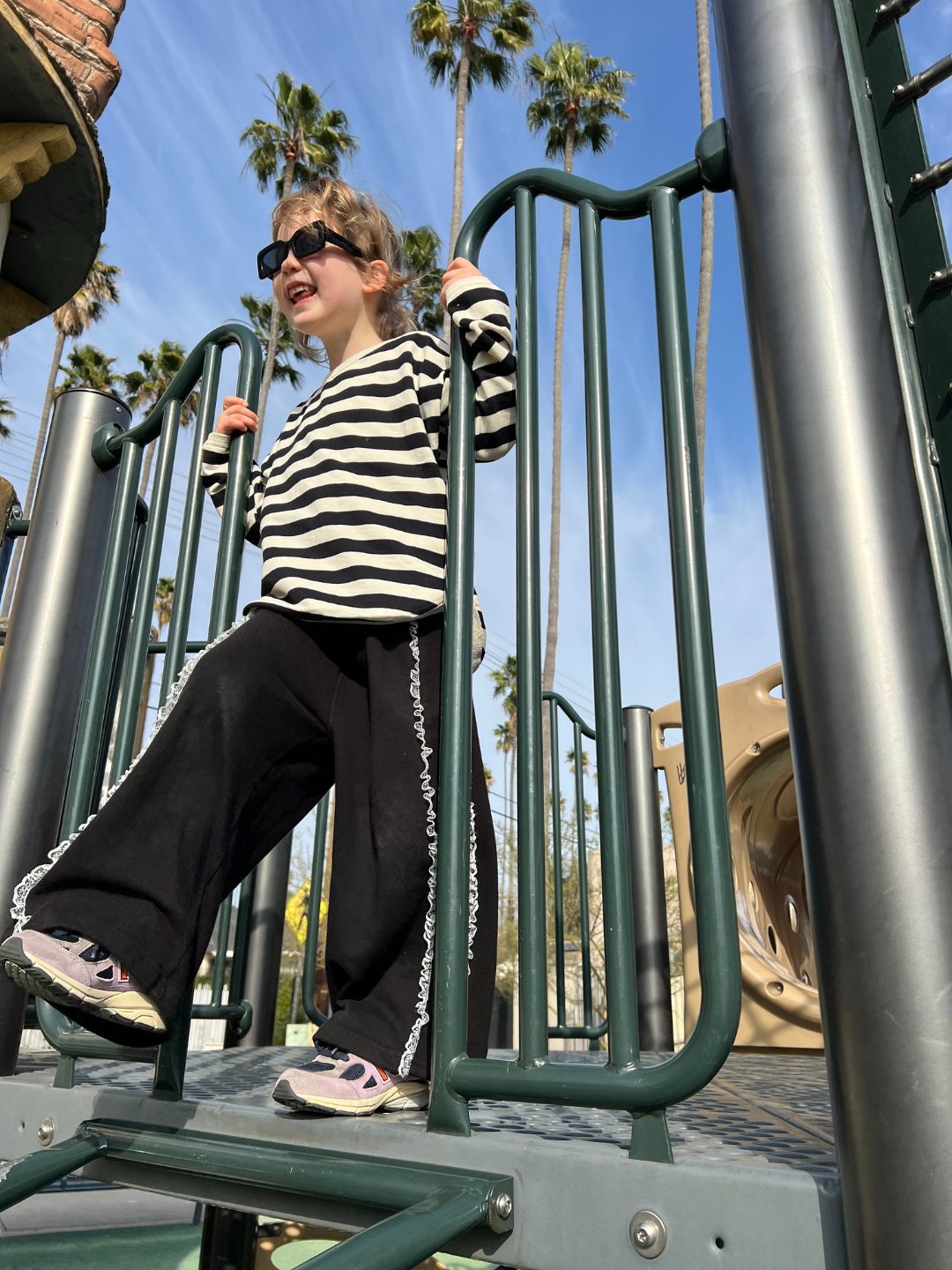 A child is wearing Relay pants with striped long sleeve tee, black sunglasses and purple sneakers