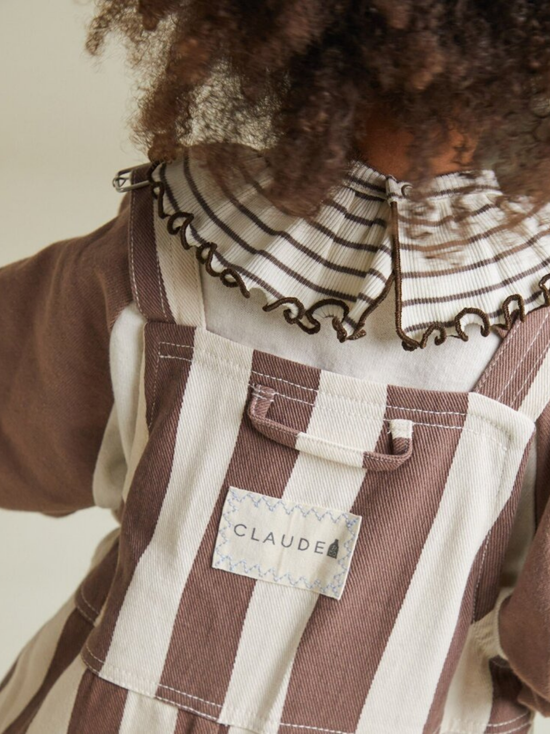 A child is shown from the back wearing BLYTHE CHOC STRIPE DUNGAREE overalls, featuring brown and white stripes, a "CLAUDE" label, and a shirt with a ruffled, striped collar.