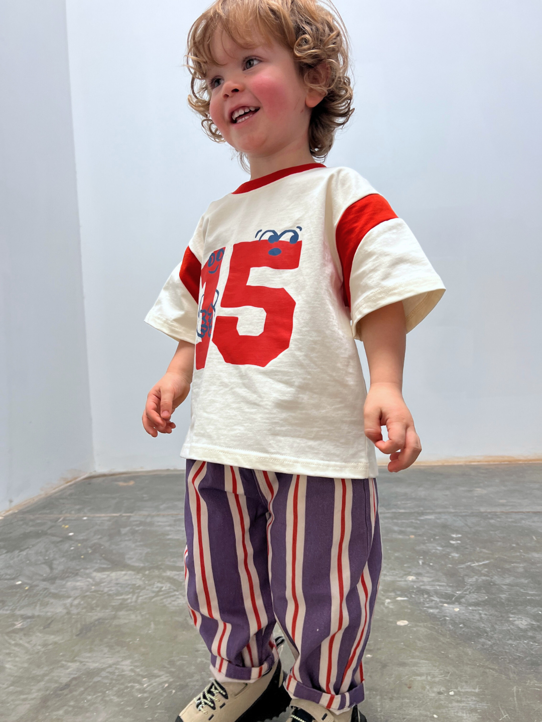 Child wearing a white shirt with red numbers and striped pants in an indoor setting