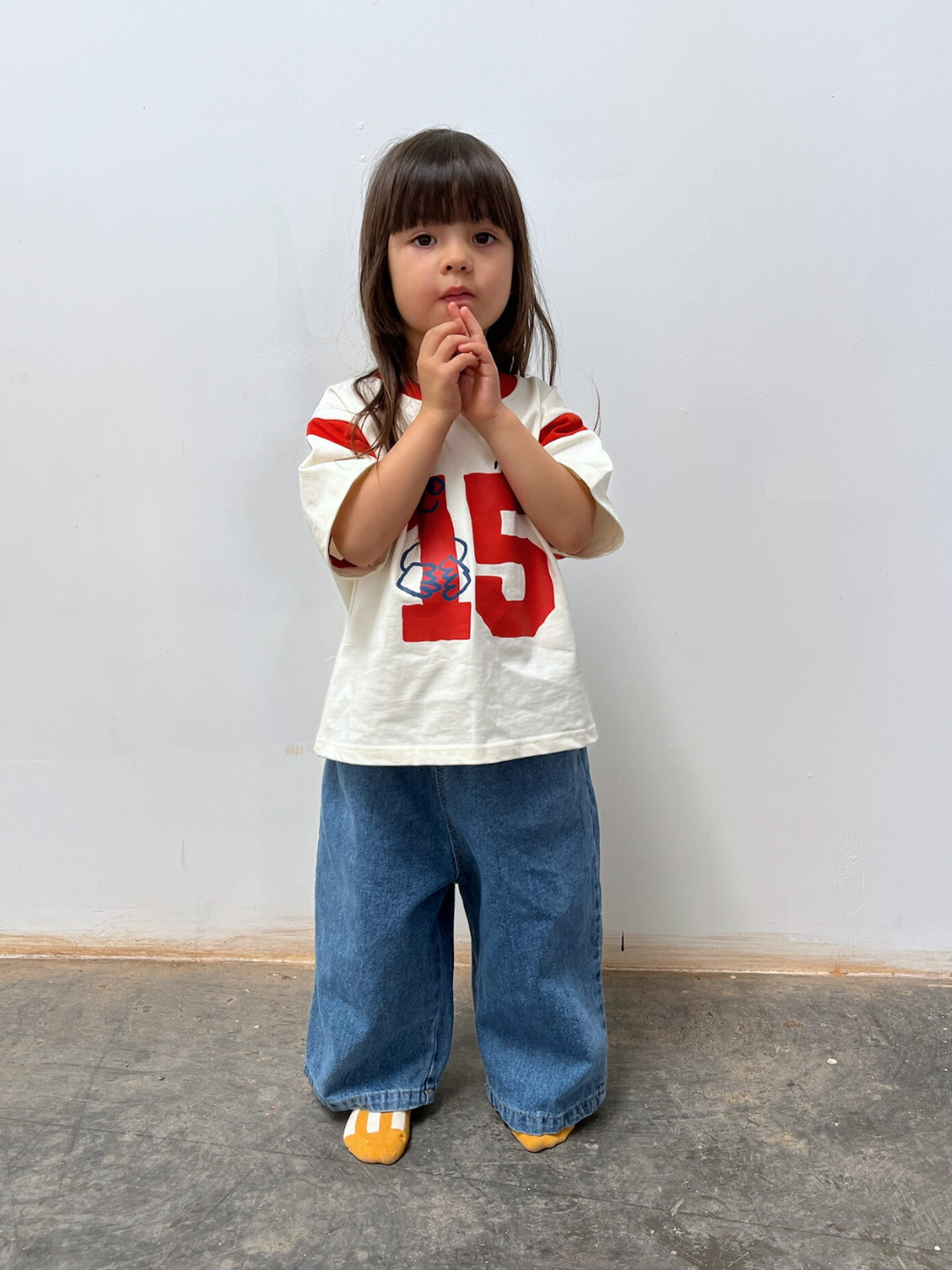 Child wearing a white t-shirt with red numbers and blue jeans on a plain background