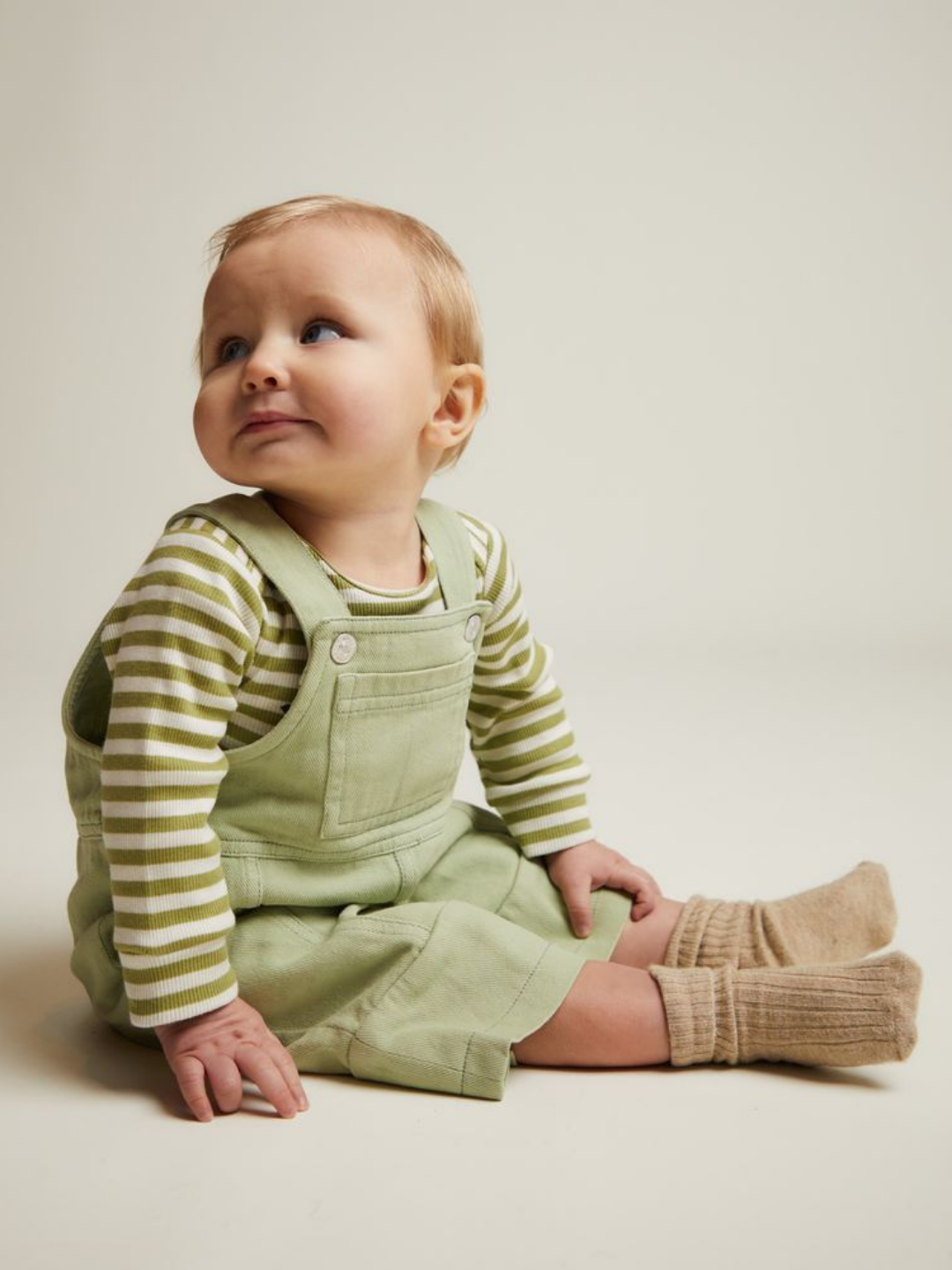 A baby wearing the BLYTHE MINT DUNGAREE with a green striped shirt and beige socks sits on the floor, gazing slightly upward.
