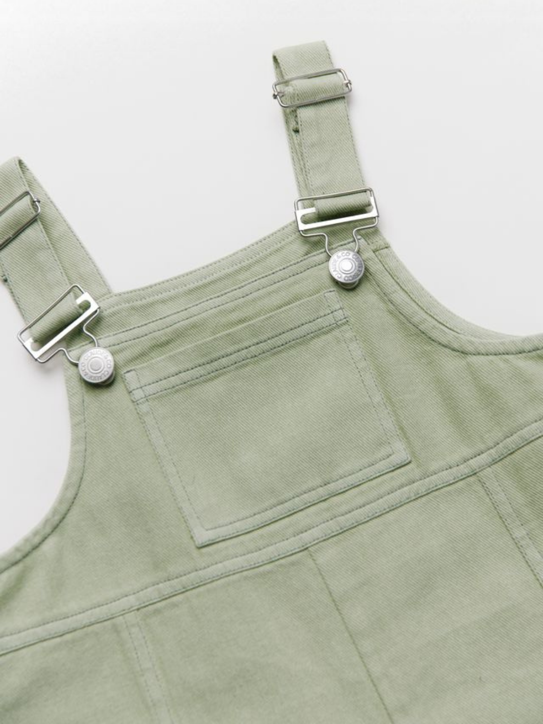 Close-up of the BLYTHE MINT DUNGAREE, a light green denim overall with a front pocket and adjustable metal buckles on the shoulder straps, laid flat on a white surface.