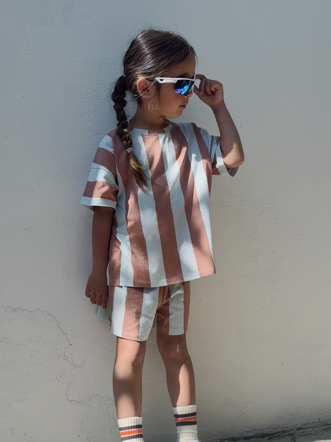 Blue/Brown | A young child in sunglasses models the UMBRELLA STRIPE SHORT SET—an organic cotton, unisex striped play set—while standing against a light-colored wall.
