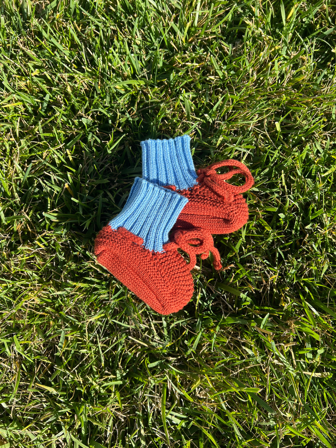 Red | Knitted baby booties in reddish orange with a blue ribbed cuff and a red tie closure, shown lying on grass