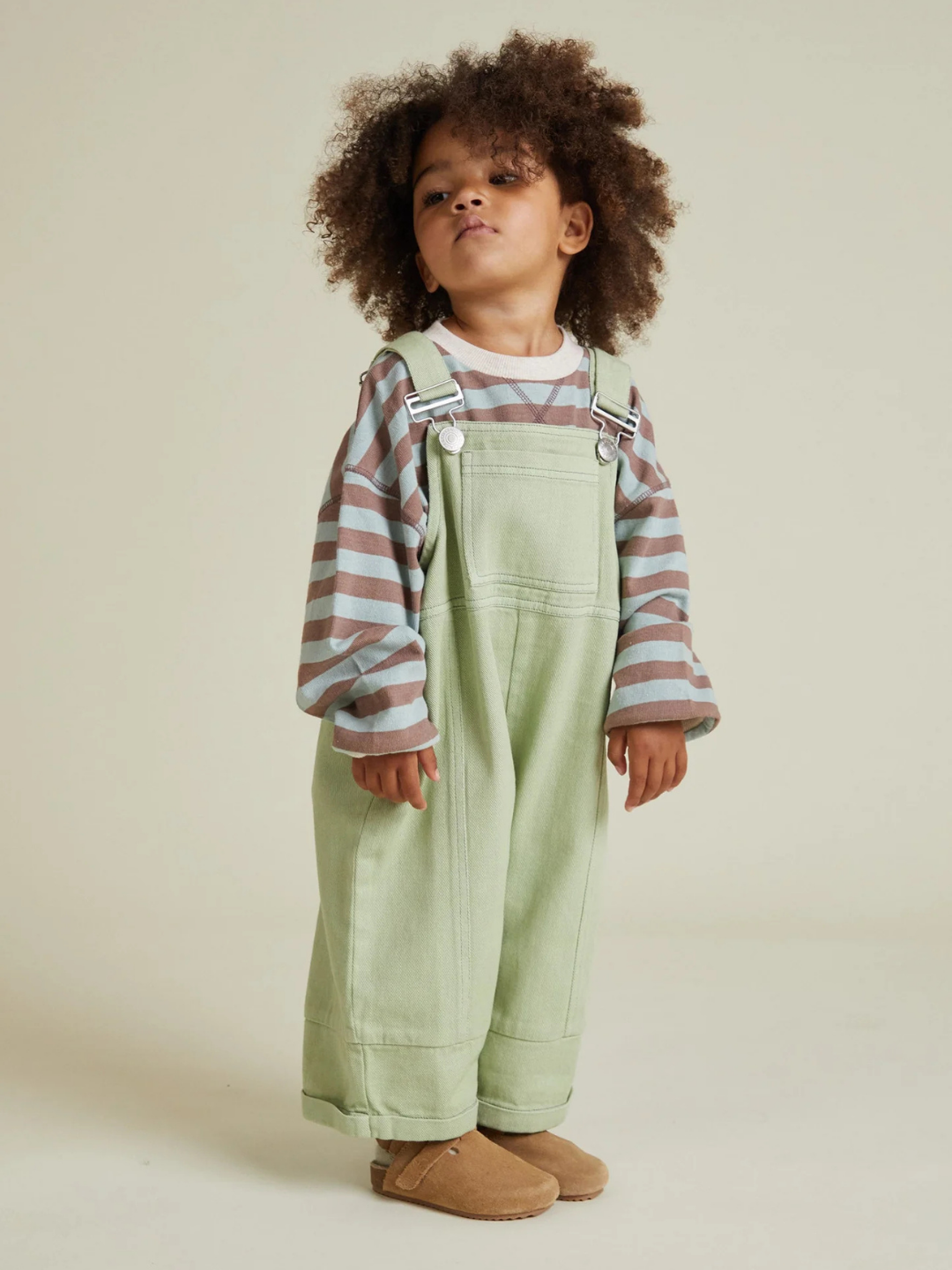 A young child with curly hair wears the BLYTHE MINT DUNGAREE over a striped long-sleeve shirt and tan slippers, standing against a plain background.