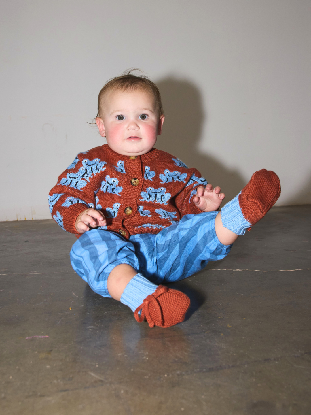 A baby sitting on a concrete floor wearing a rust-red cardigan with light blue cartoon frog patterns, matching blue striped pants, and rust-brown booties.