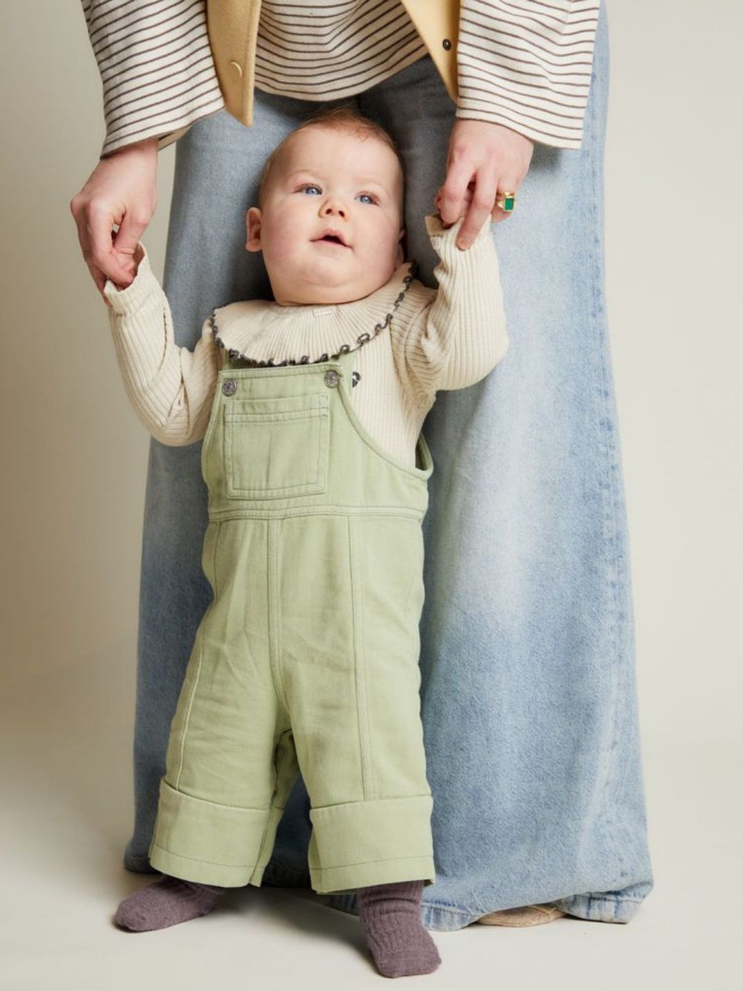 An adult helps a baby stand by holding their hands. The baby wears the BLYTHE MINT DUNGAREE with a cream shirt, while the adult wears wide-legged jeans and a striped top.