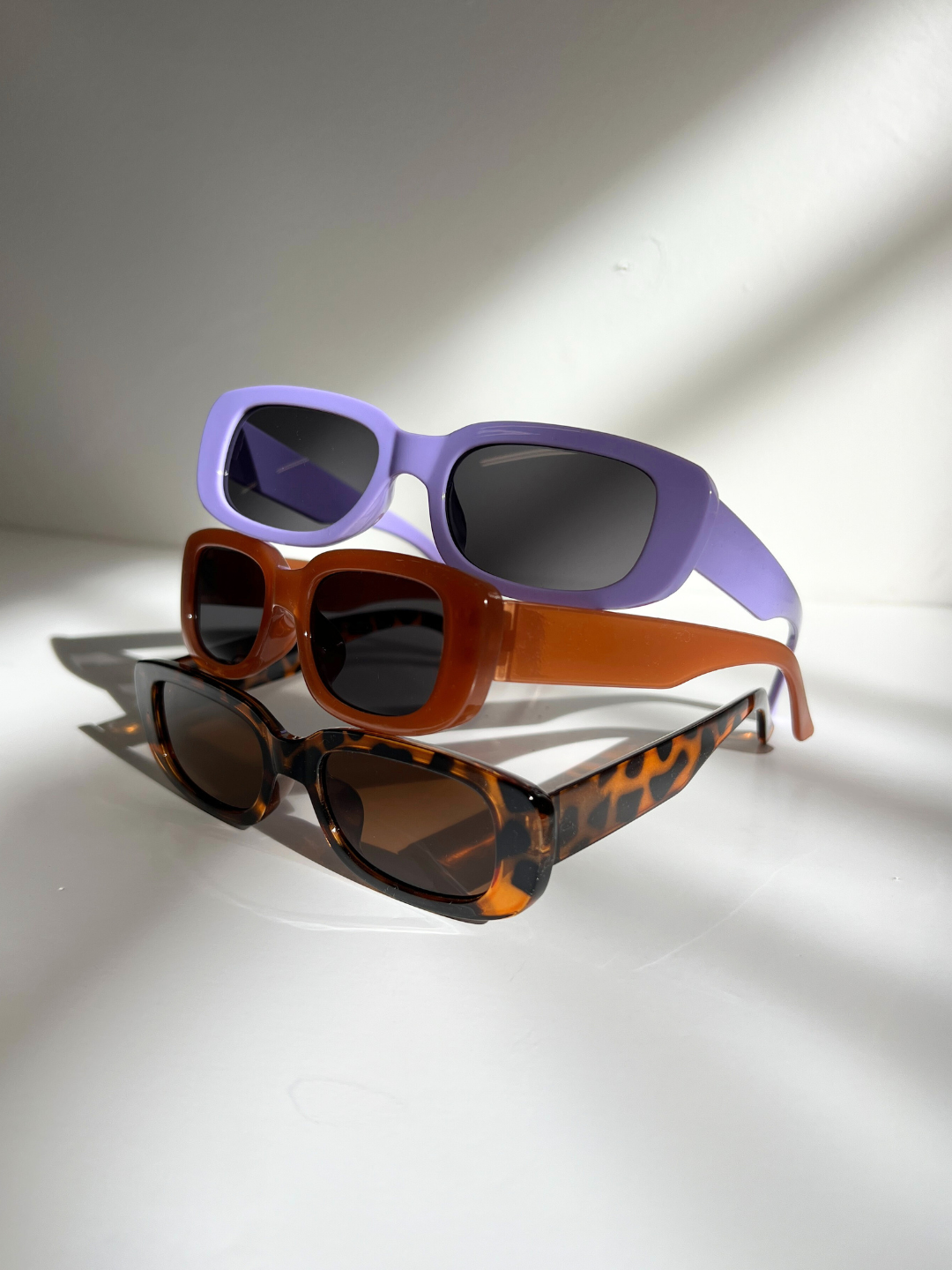 Tortoiseshell | Three pairs of SANTA CRUZ SUNGLASSES, ideal for ages 2-6, rest on a white surface. They come in purple, orange, and tortoiseshell patterns and feature UV400 lenses for maximum eye protection. Light creatively casts shadows in the background.