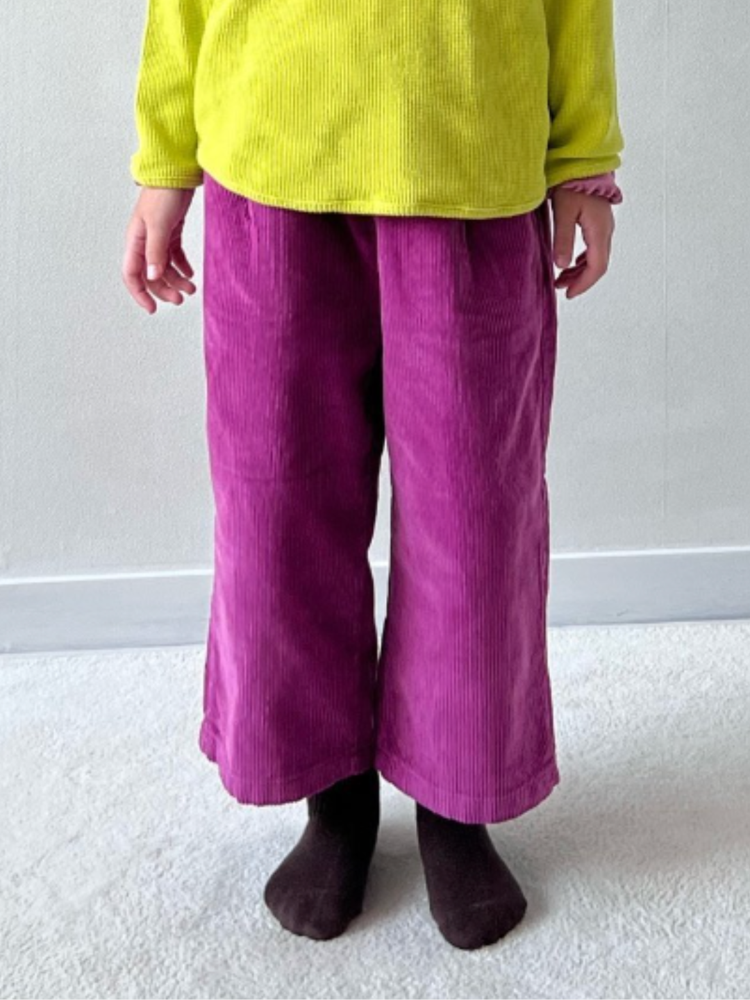 Magenta | Waist-down cropped image of a child wearing wide-leg corduroy pants in magenta pink, paired with a yellow shirt and dark brown socks, standing on grey carpet..