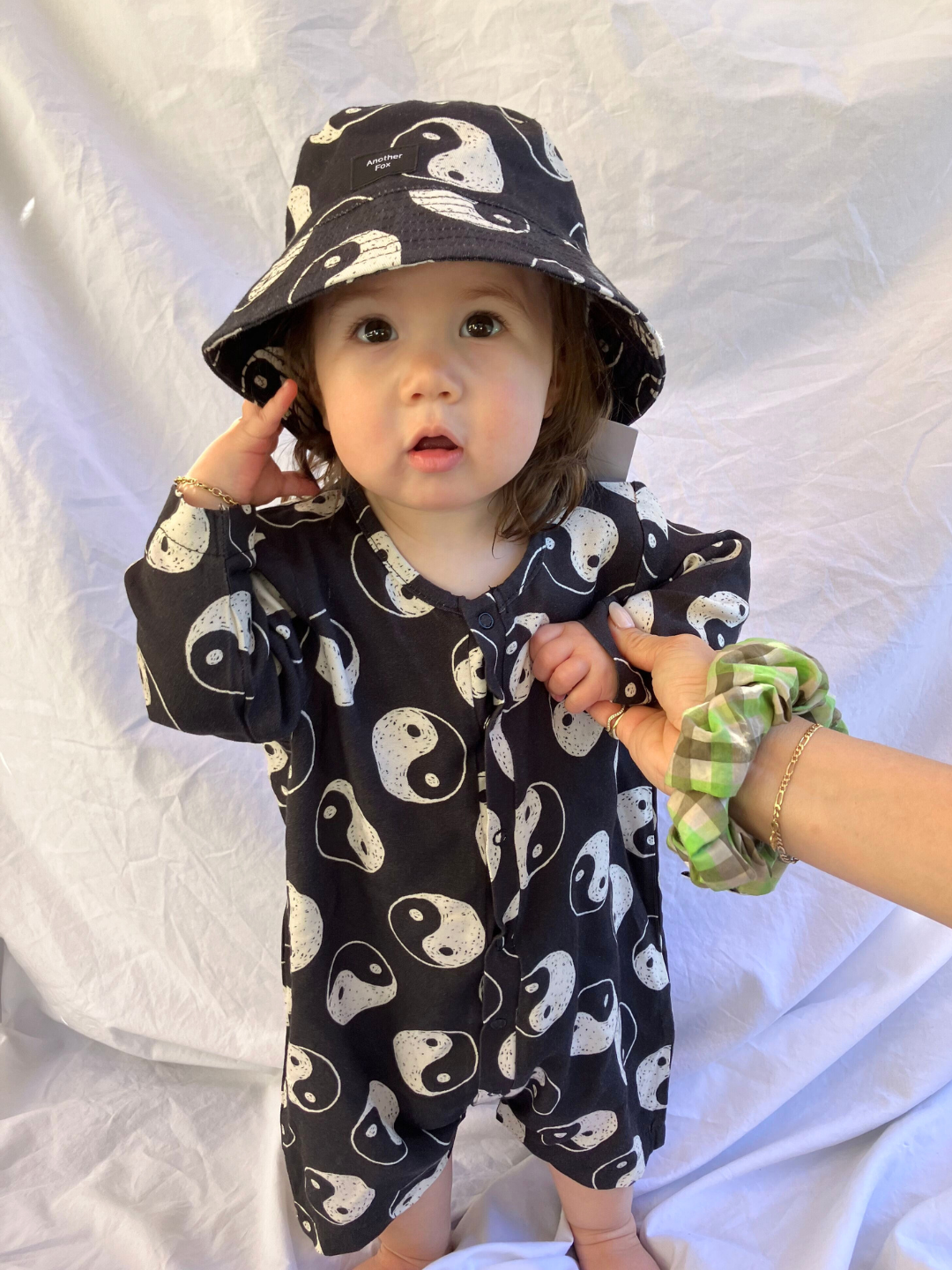 A toddler wearing the yin yang bucket hat with a matching short jumpsuit in black cotton printed all over white white yin yangs. She holds an adults hand with a green plaid scrunchie on the wrist, and is standing on a white sheet backdrop.