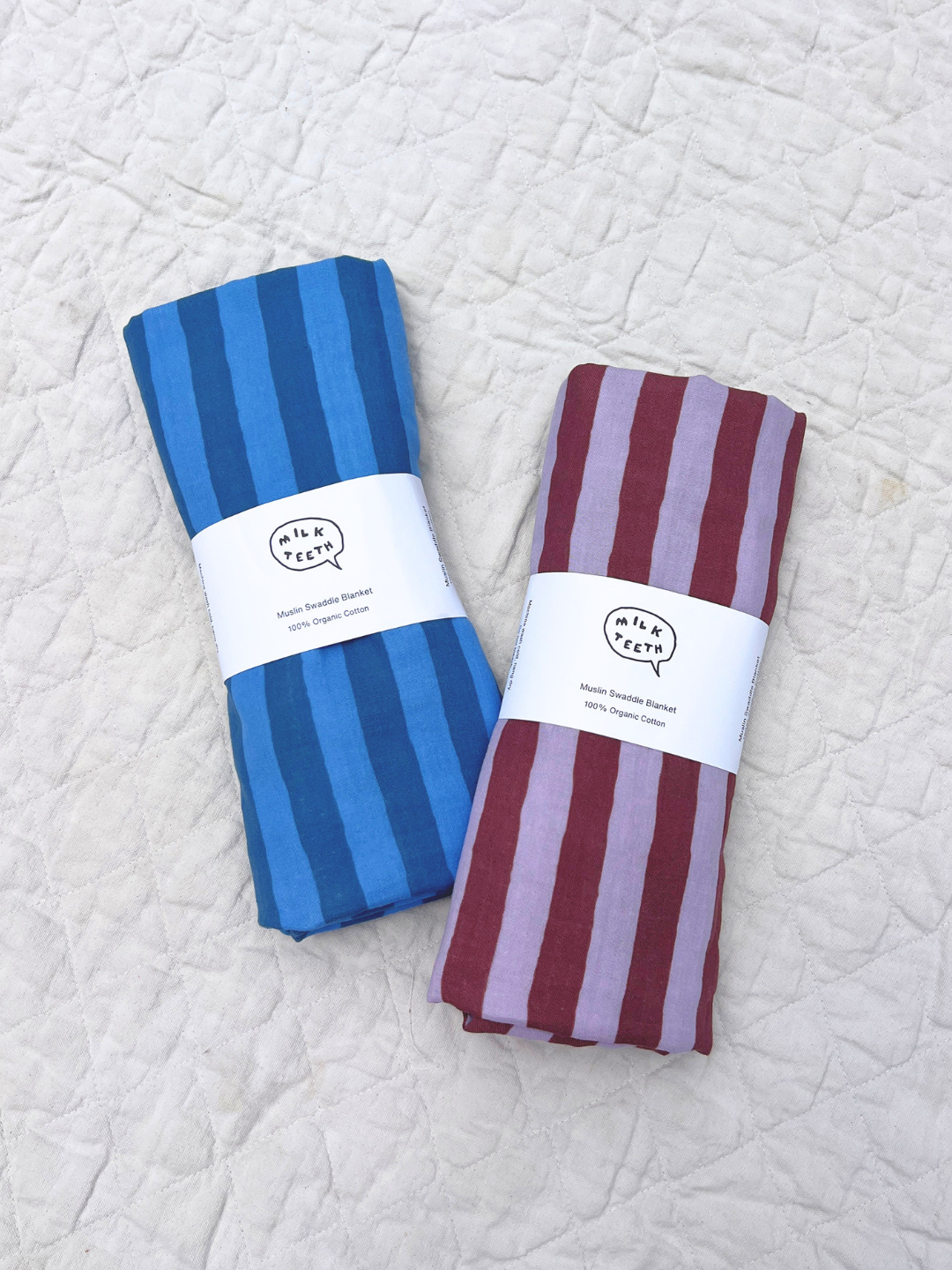  Two organic cotton swaddles, one in blue stripes and one in purple and maroon stripes are shown rolled up with a white Milk Teeth label around the of each, and laid on a beige quilt.
