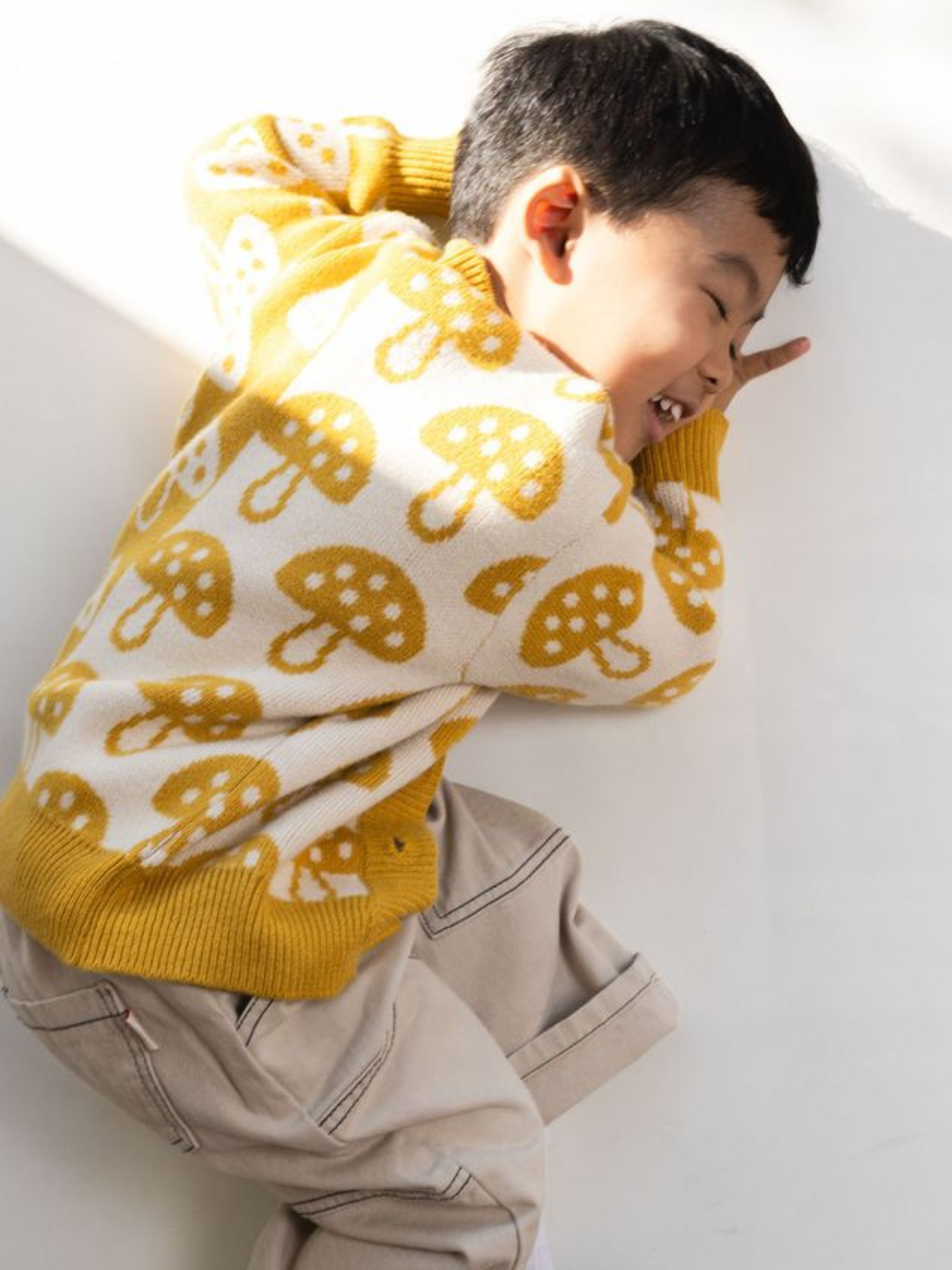 A young child wearing the SHROOM CARDIGAN, a yellow sweater adorned with mushroom patterns, paired with beige pants, lies on their side, smiling and covering their face with one hand, perfectly capturing the joy of childhood in this vivid description.