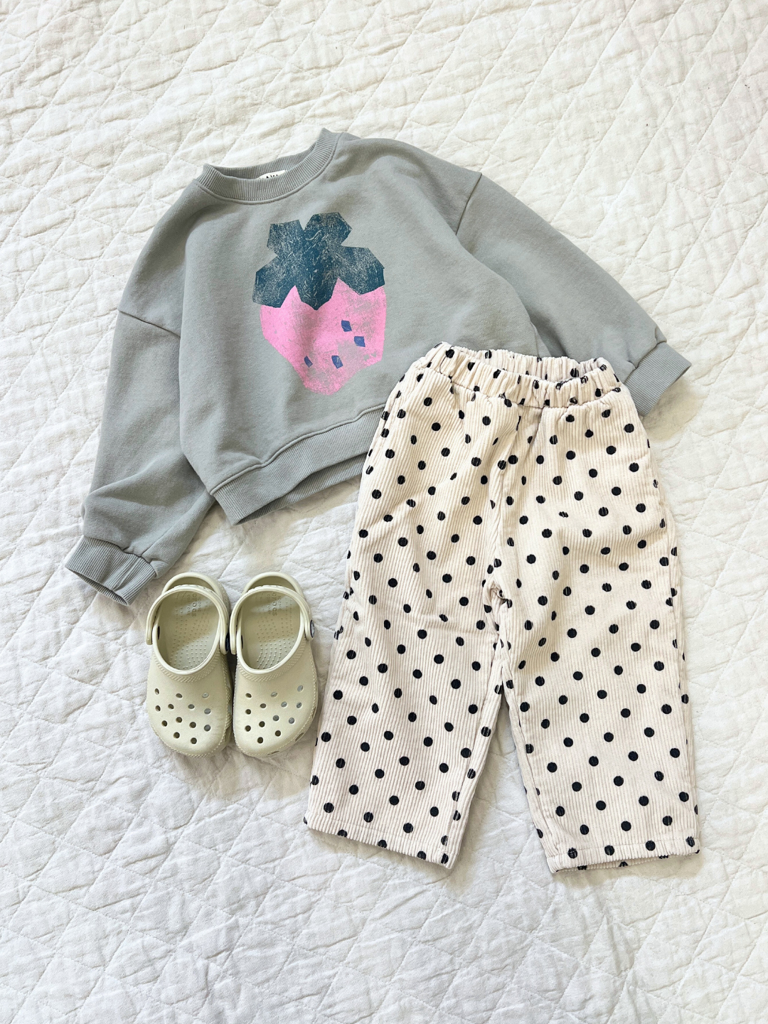 Children's outfit includes a STRAWBERRY CROPPED SWEATSHIRT, cream polka dot pants, and light green slip-on shoes displayed on a quilted surface.