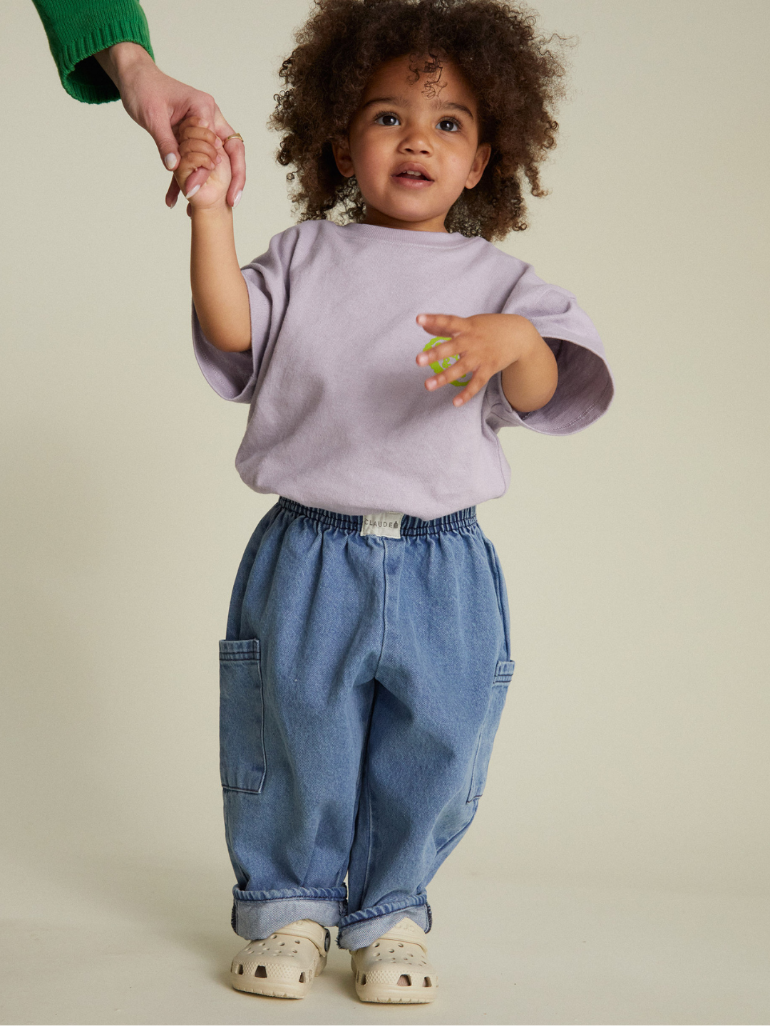 A young child with curly hair, wearing a lavender shirt, EMERSON DENIM BABY TROUSER, and white sandals, holds an adult's hand against a plain background.