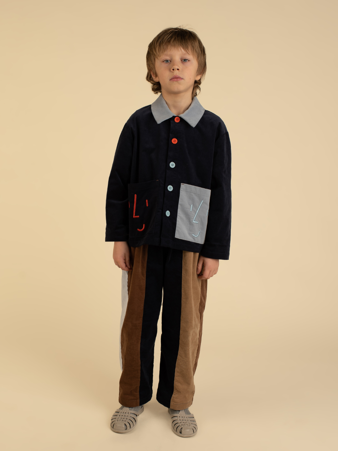 Navy Blue | A child models the FACE JACKET in navy, brown, and gray with colorful buttons and pocket accents, standing against a beige background and looking at the camera.