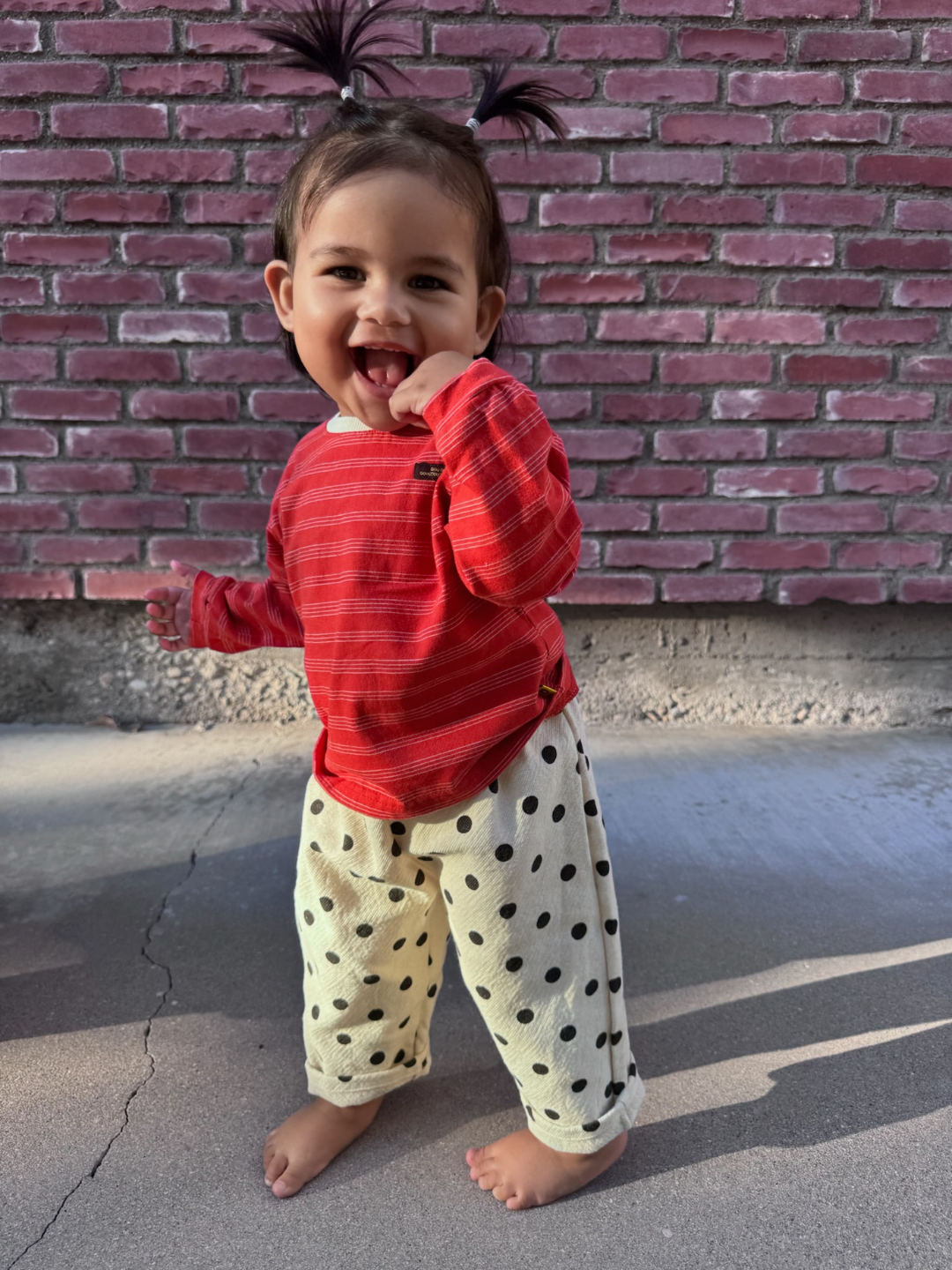 Child wearing a red striped shirt and polka dot pants standing in front of a brick wall.
