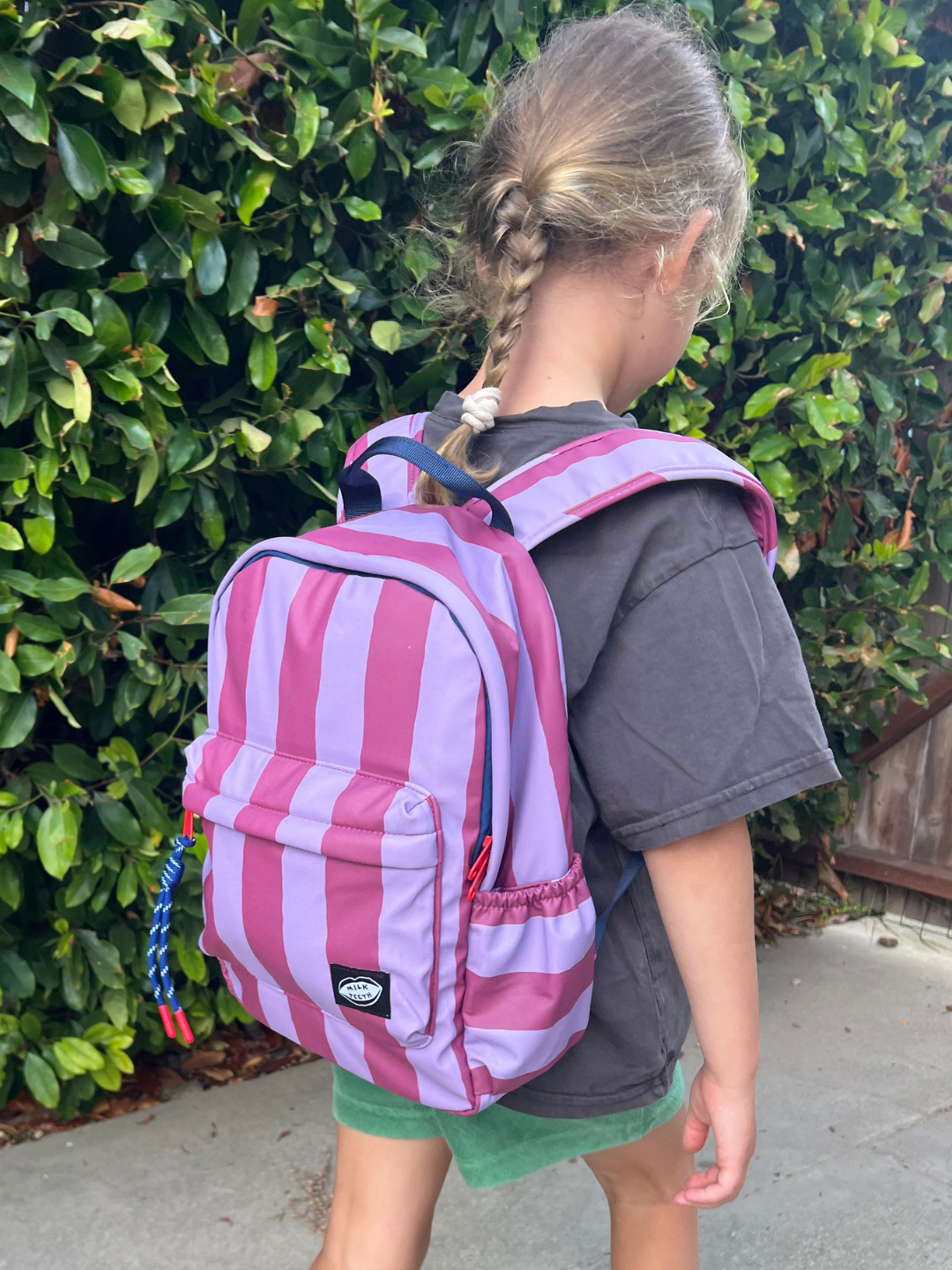 Purple Stripe | A child wearing a maroon and lavender striped backpack, standing outdoors in front of a green hedge.