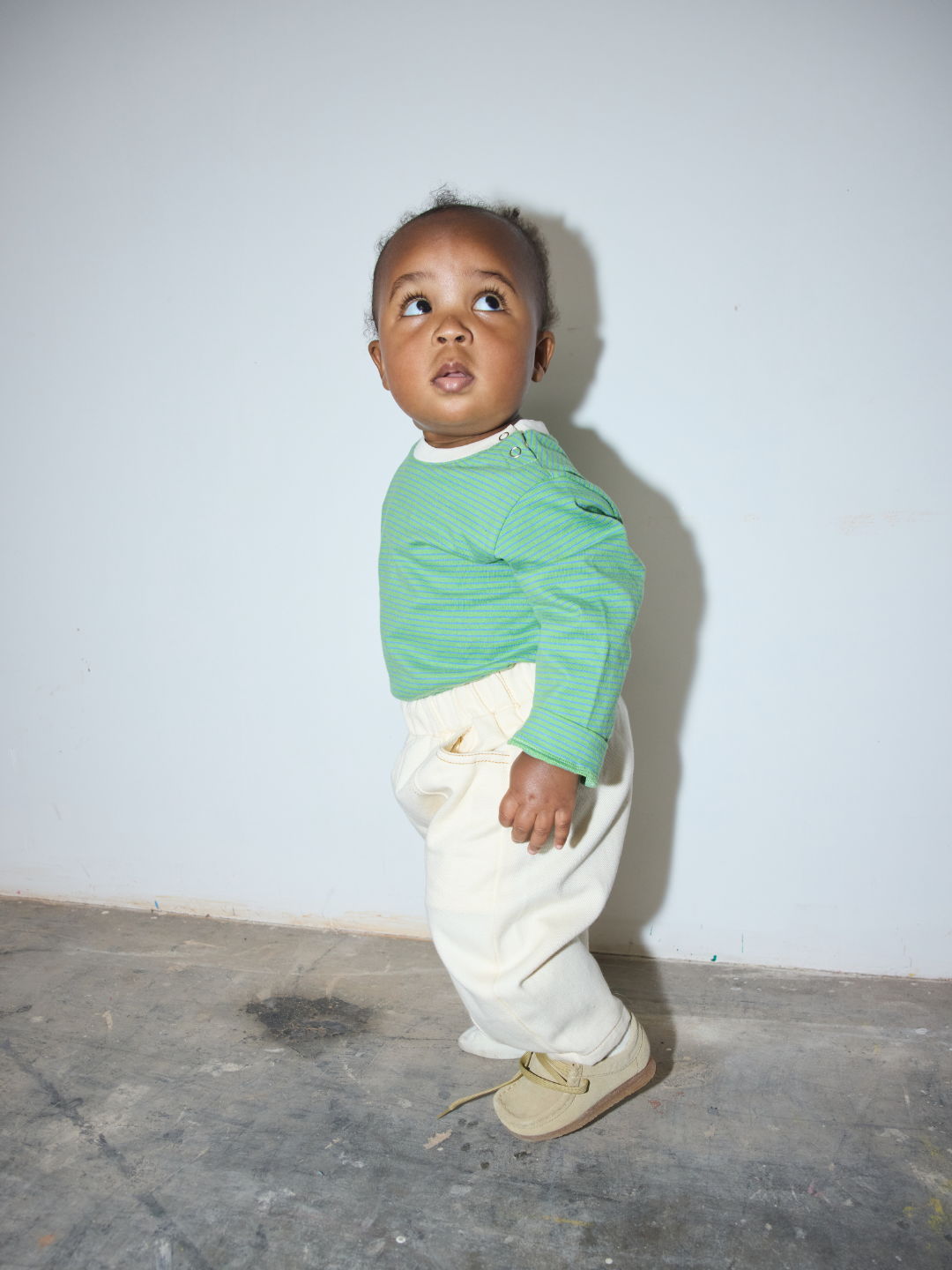 Green/Blue | A toddler standing against a plain light gray wall on a concrete floor, wearing a green long-sleeve top, cream pants, and tan shoes.
