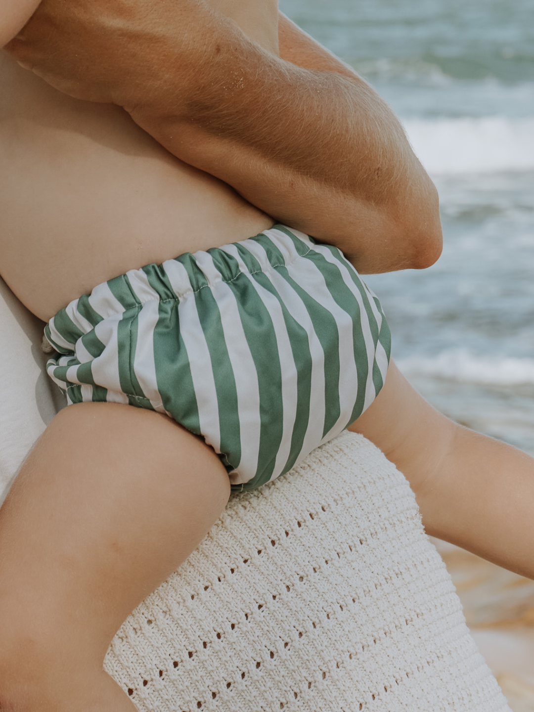The back view of the swim diaper with an elastic waist with a tie and elastic leg holes on a child being held by their parent at the beach. The diaper is a has cream and moss green vertical stripes.