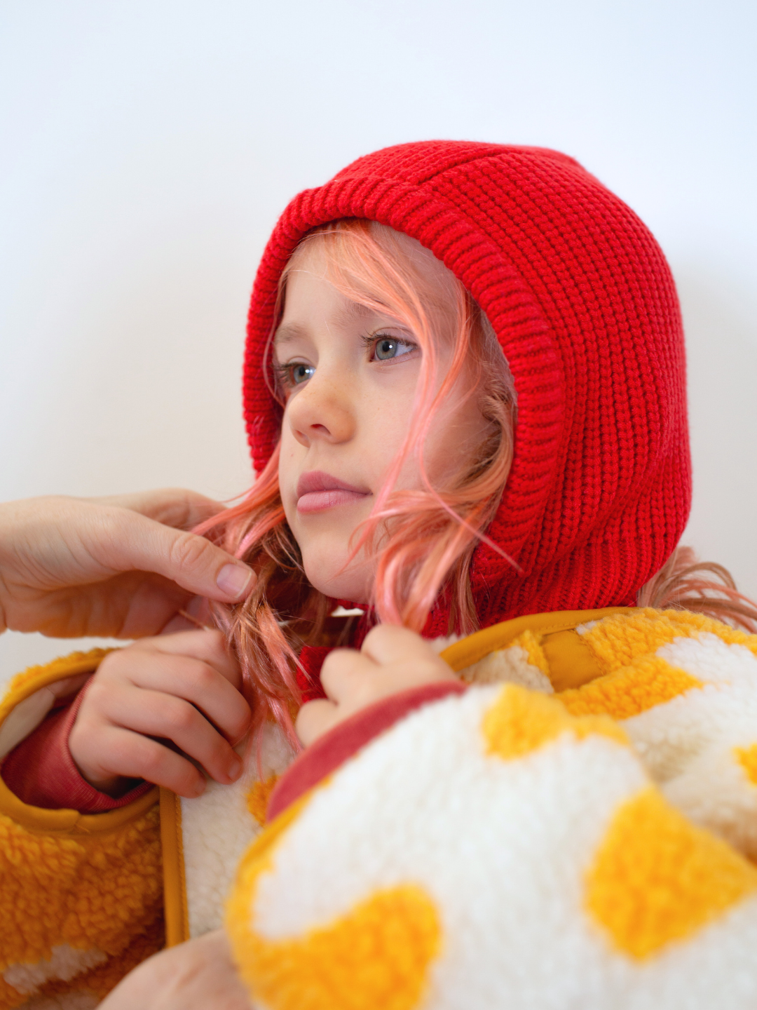 Child wearing a red knit hat and yellow and white patterned garment against a light background