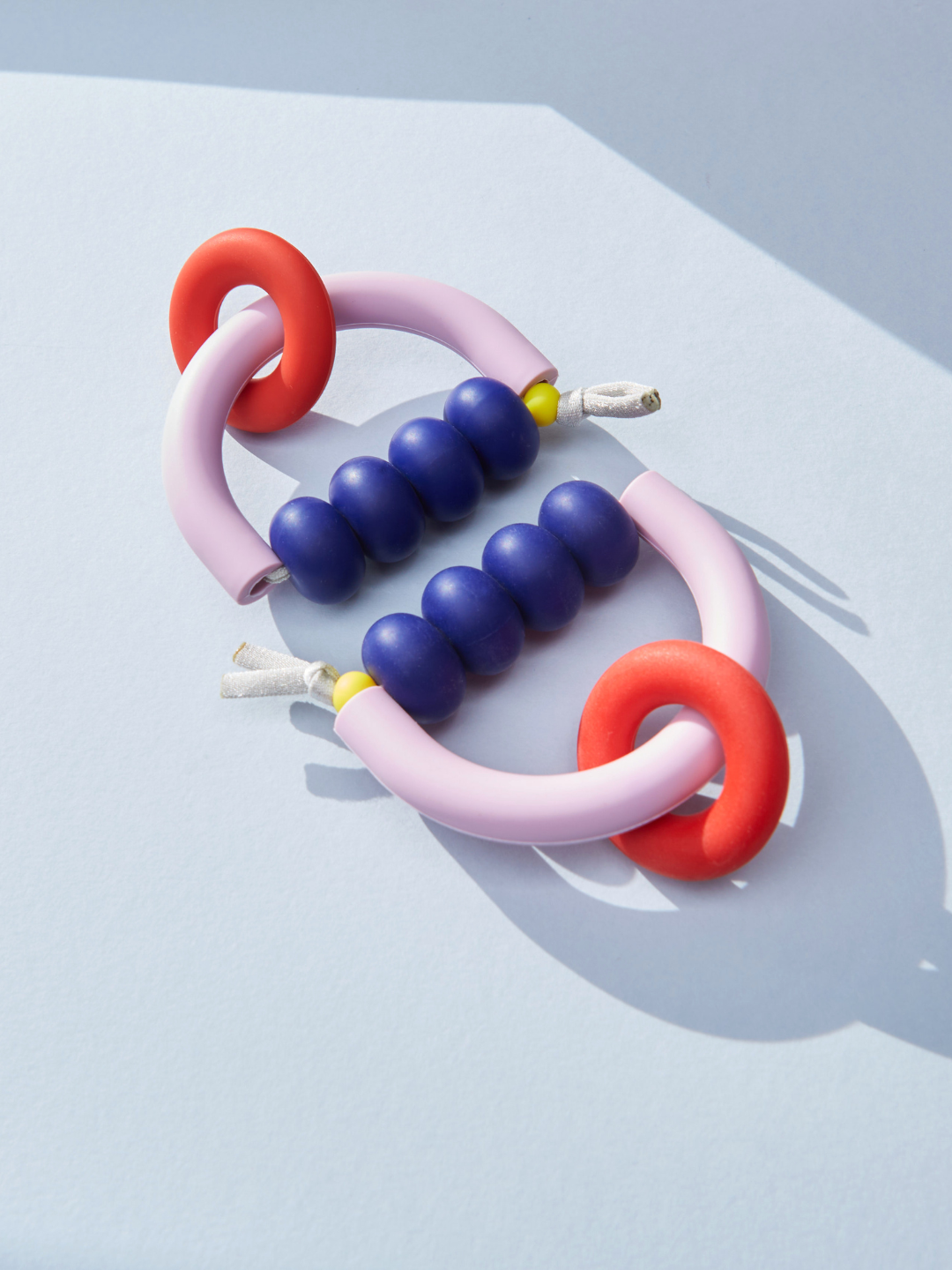 Sunset | Two pastel pink loops with purple beads and red rings from the MT X JM ARCH RING TEETHER, a US-made sensory-friendly teether, are arranged on a light blue surface with soft shadows.