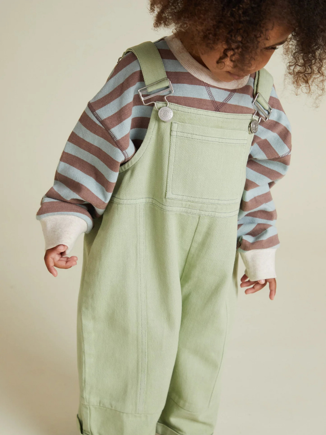 Child wearing BLYTHE MINT DUNGAREE and a long-sleeve shirt with brown, blue, and white stripes, standing against a plain background.