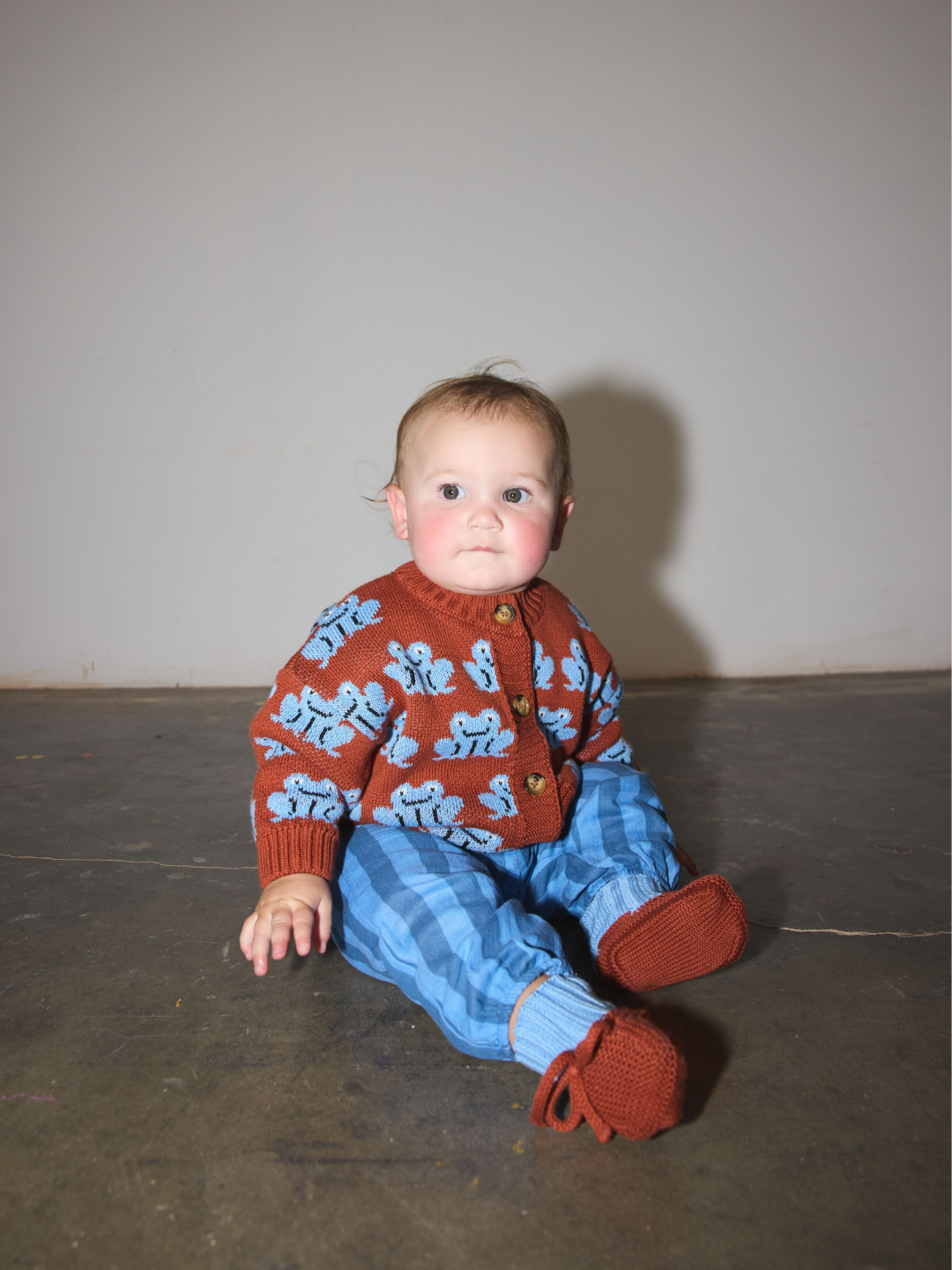 Red | A baby sitting on a concrete floor wearing a rust-red cardigan with light blue cartoon frog patterns, matching blue striped pants, and rust-brown booties.
