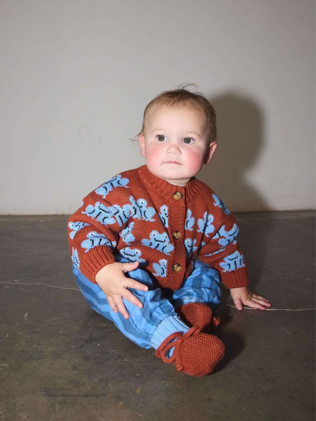 A baby sitting on a concrete floor wearing a rust-red cardigan with light blue cartoon frog patterns, matching blue striped pants, and rust-brown booties.