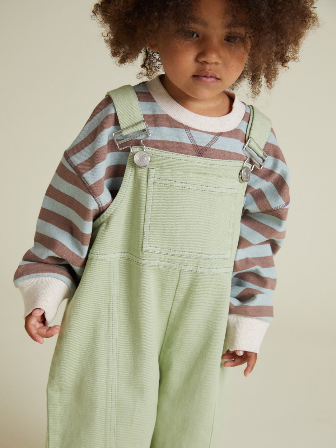A child in the BLYTHE MINT DUNGAREE and a brown and blue striped long-sleeve shirt stands against a plain, light background.