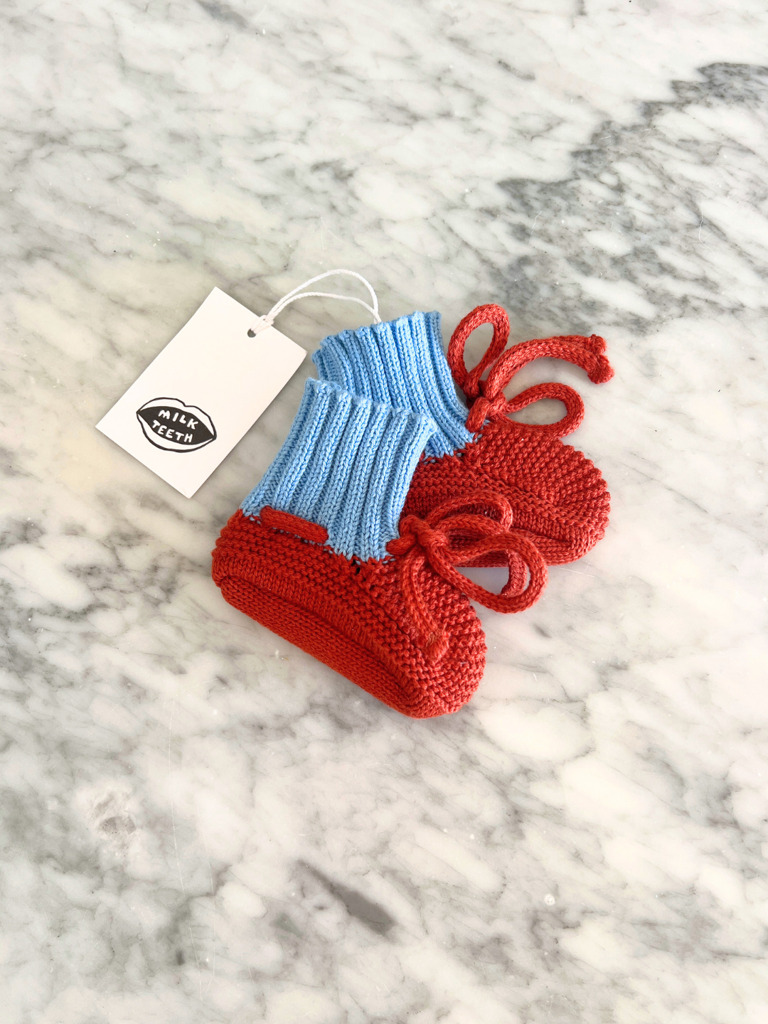 Red | Knitted baby booties in reddish orange with a blue ribbed cuff and a red tie closure and a Milk Teeth logo tag, shown lying on a grey marble surface