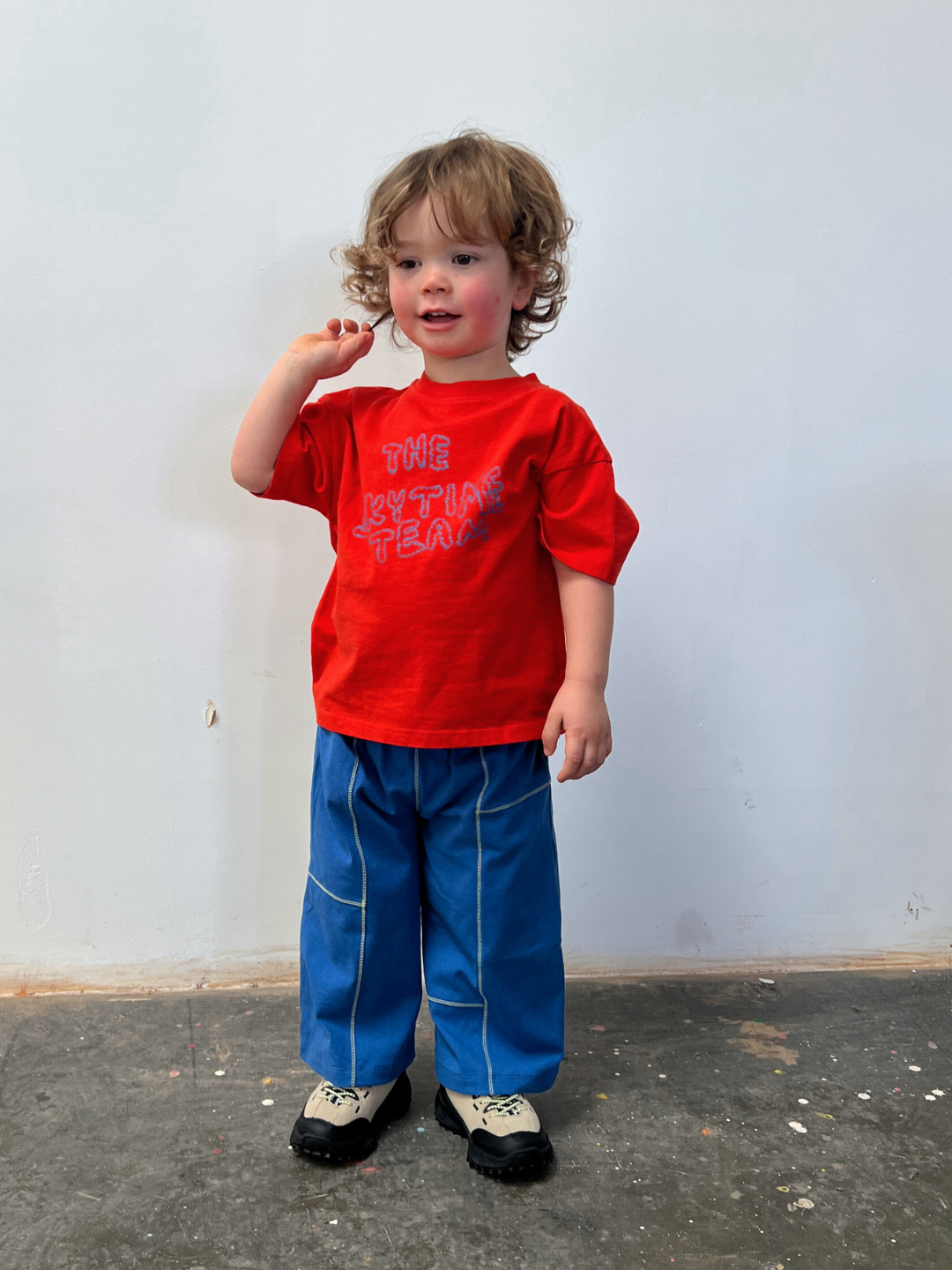 Child wearing a red shirt and blue pants standing against a plain wall.