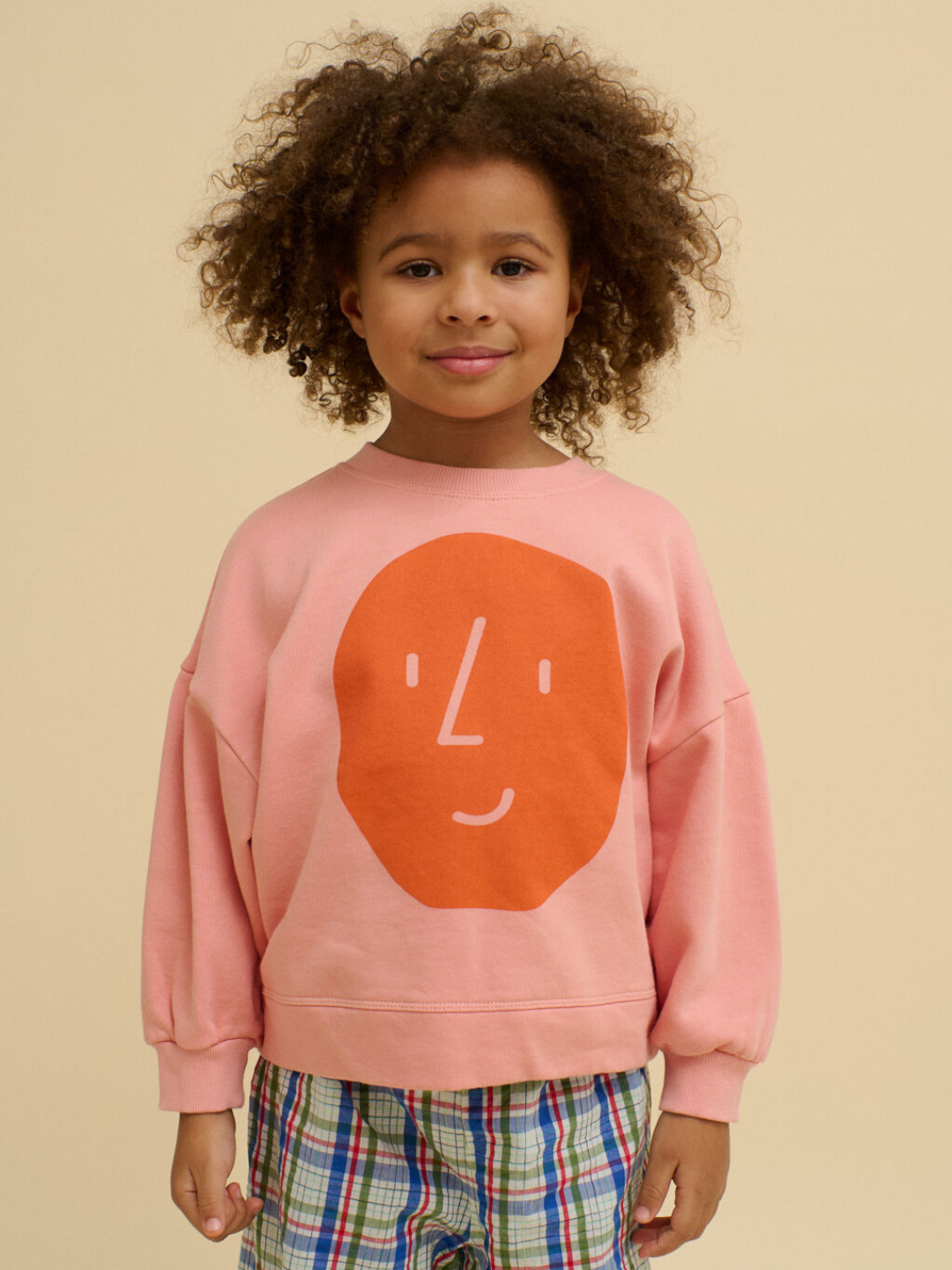 A child with curly hair wears a BOXY POTATO SWEATSHIRT—an oversized organic cotton top in pink with an orange smiley face—paired with plaid shorts, standing against a beige background.