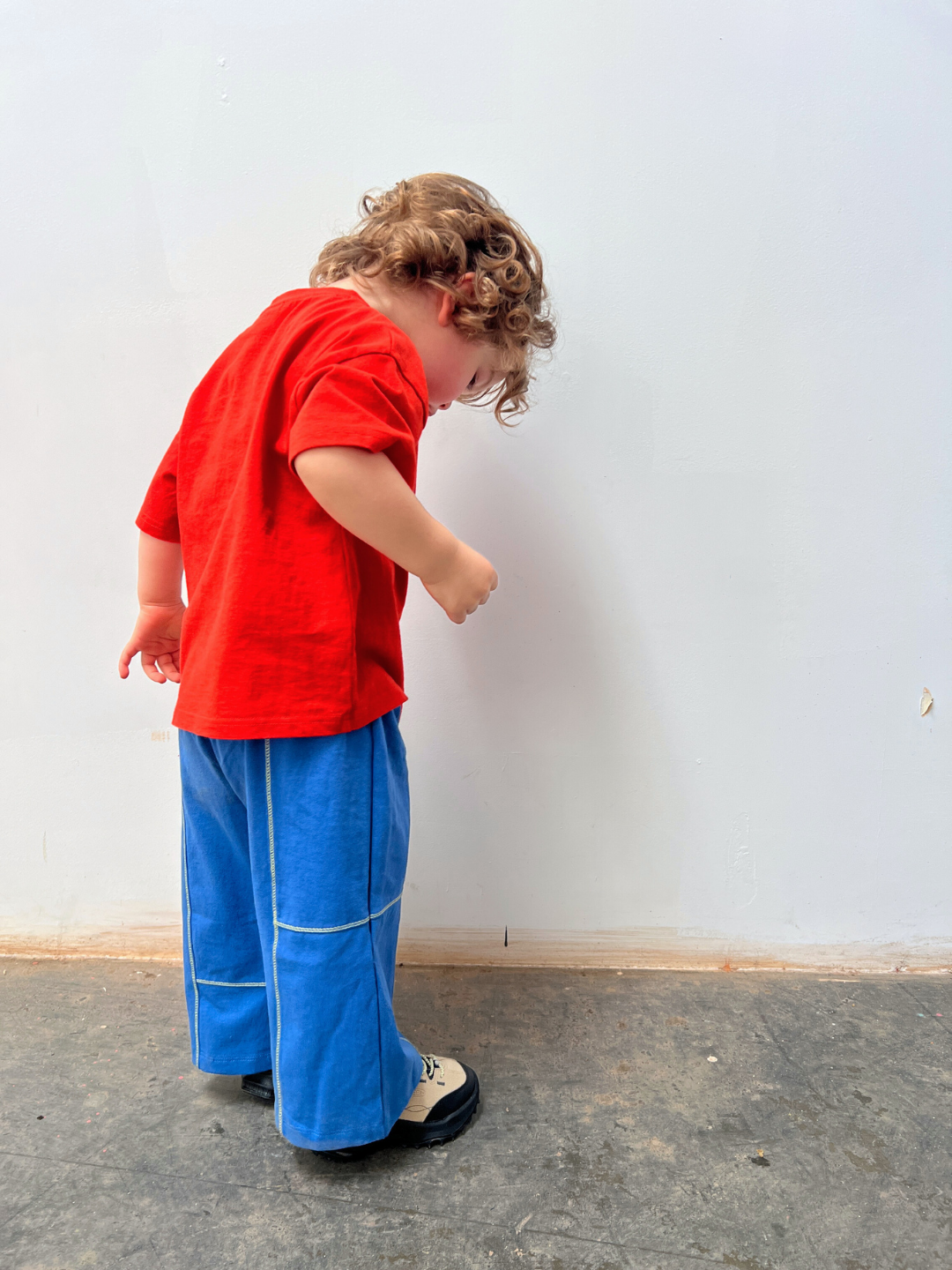 Red | Child wearing a red shirt and blue pants standing against a white wall.