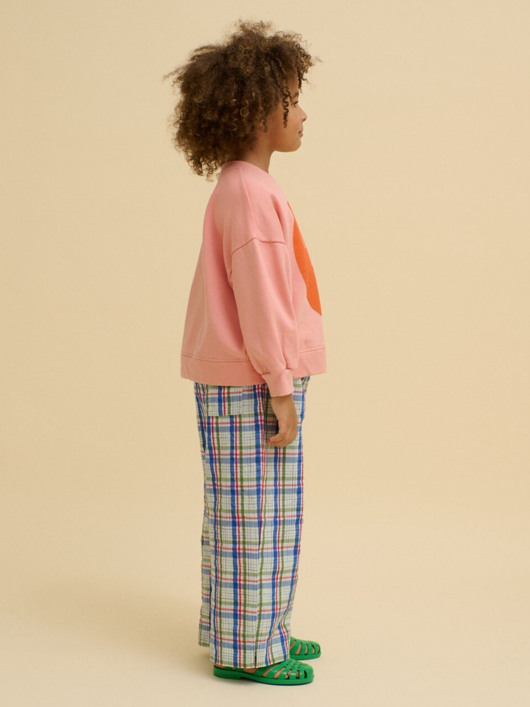 A child with curly hair stands in profile, wearing a pink BOXY POTATO SWEATSHIRT, plaid pants, and green sandals—all made from soft organic cotton—against a beige background.