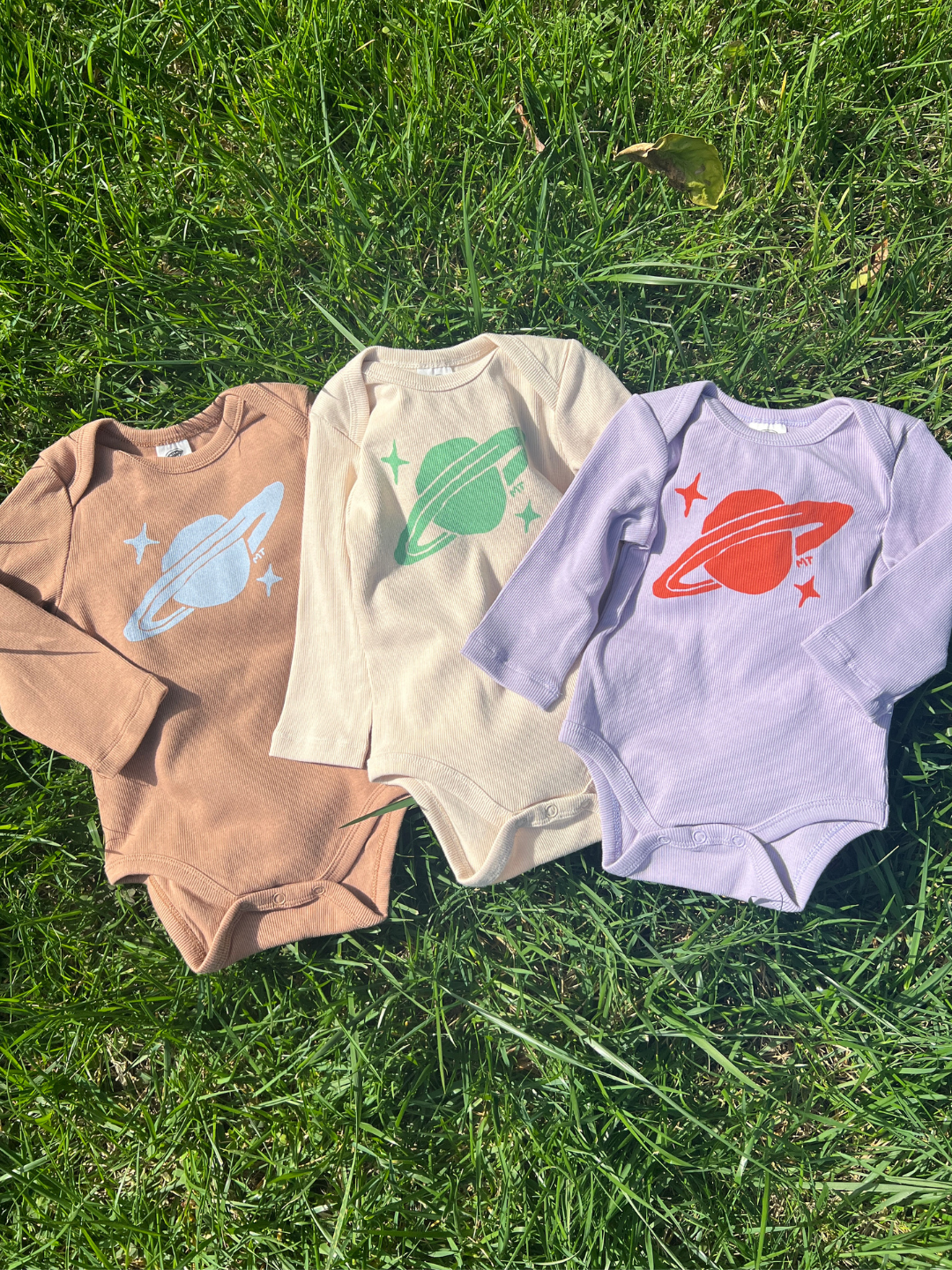 Three SATURN LONG SLEEVE ONESIES in brown, cream, and lavender with snaps for easy diaper changes are laid on grass; each features a ribbed cotton design inspired by planets.