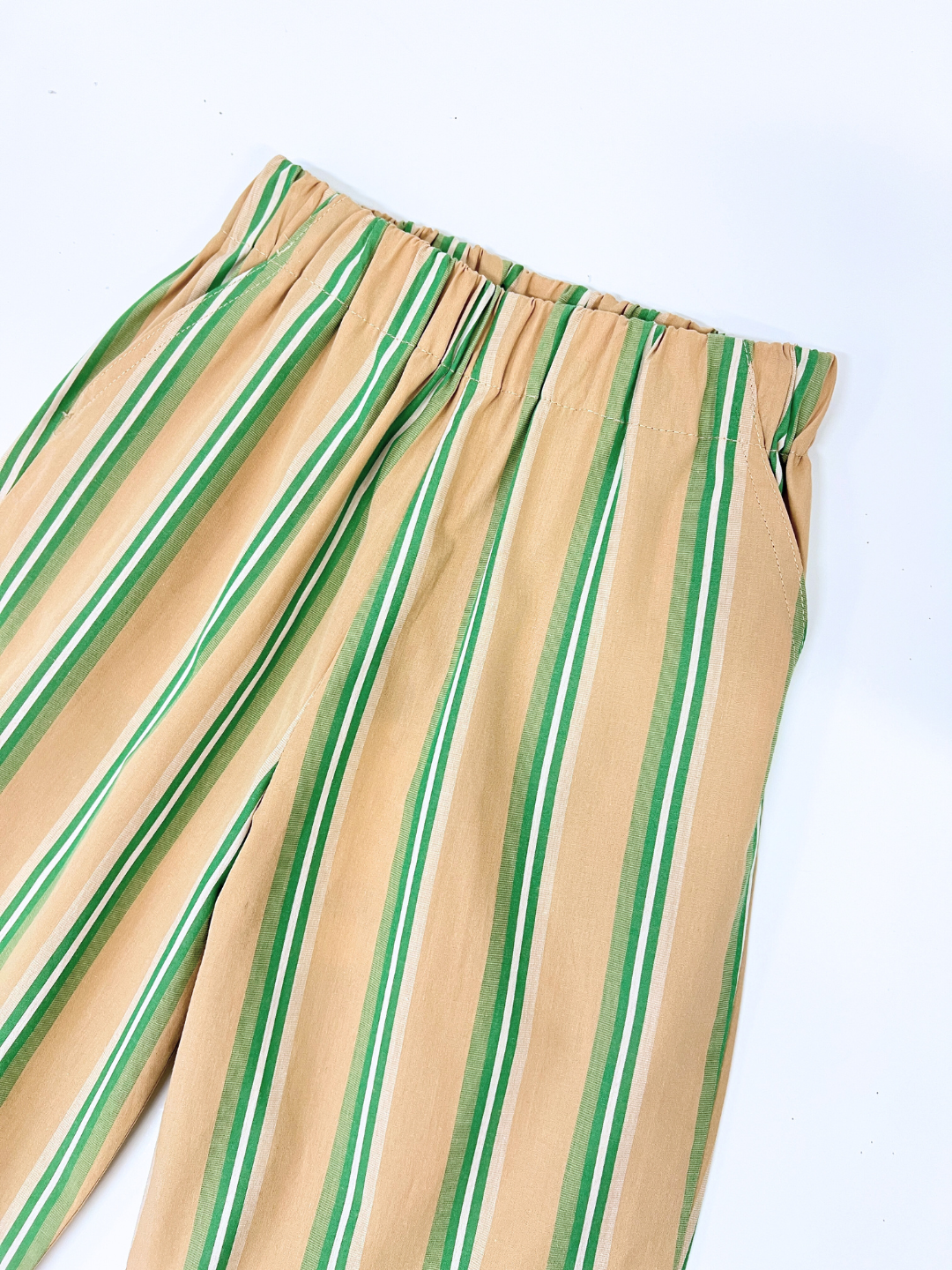 The EVER WIDE-LEG STRAIGHT STRIPED TROUSER is a unisex children's pant with tan fabric, green and white vertical stripes, an elastic waistband, and side pockets. Made from organic cotton for comfort and style on a white background, perfect for sustainable kidswear collections.