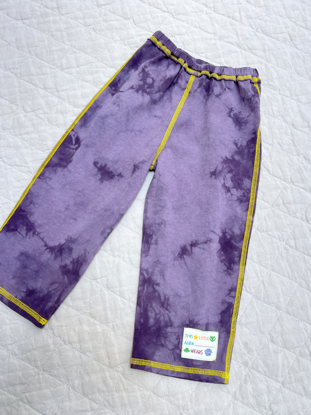 A pair of COSMO TIE DYE PANT in purple cotton jersey with yellow stitching and a "This Little Alien Wears" label, laid flat on a light quilted surface.