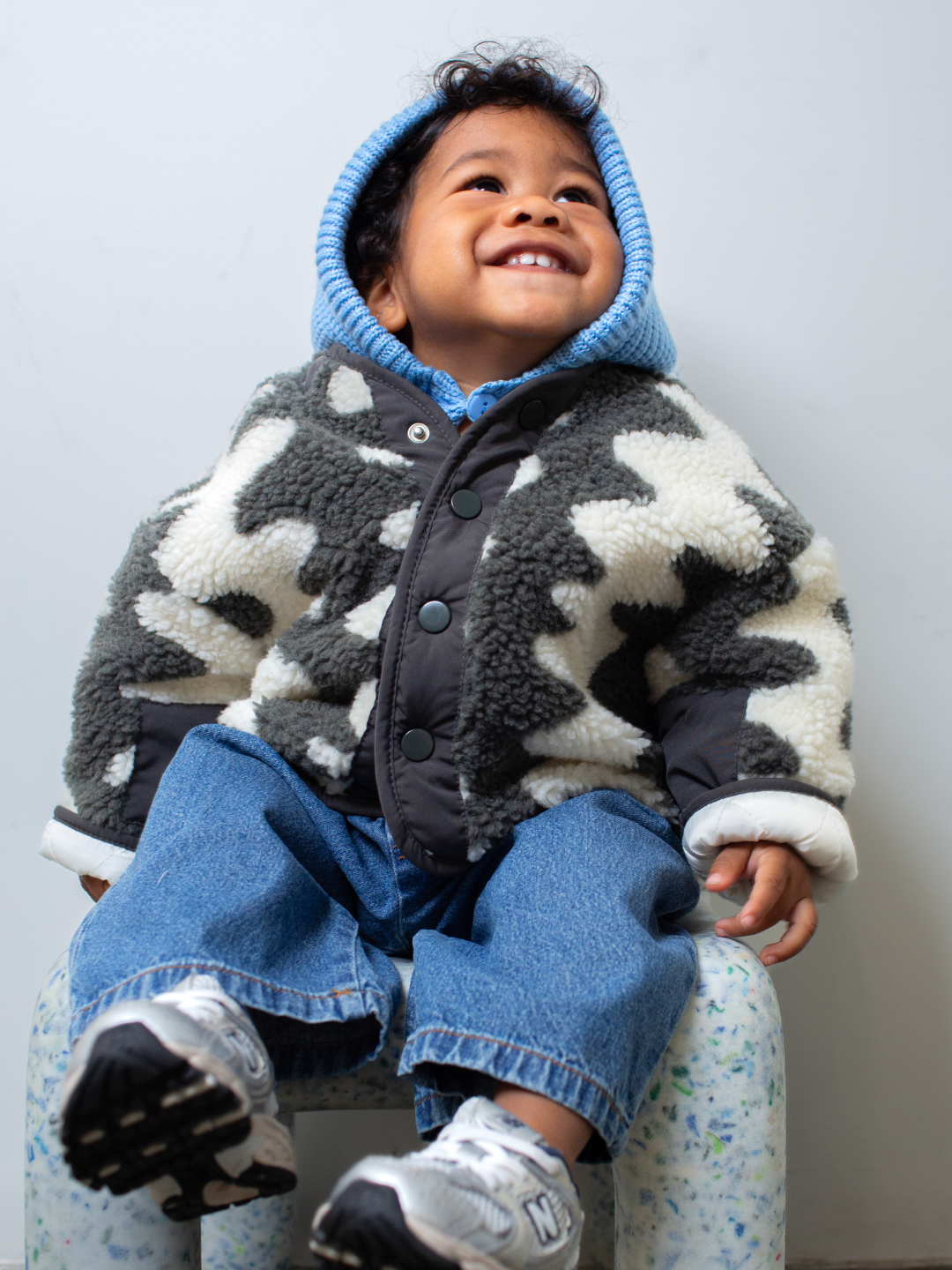 Child sitting on a small stool wearing a patterned jacket, sky blue hood and blue jeans against a plain background